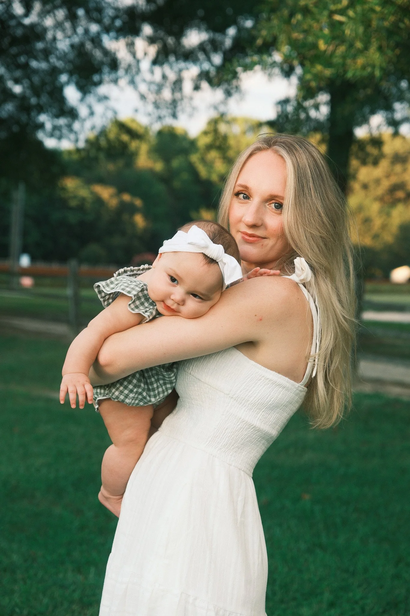 A woman with long blonde hair holding a baby girl outdoors. The woman is wearing a white dress, and the baby is dressed in a green checkered outfit with a white headband. Trees and greenery are in the background.