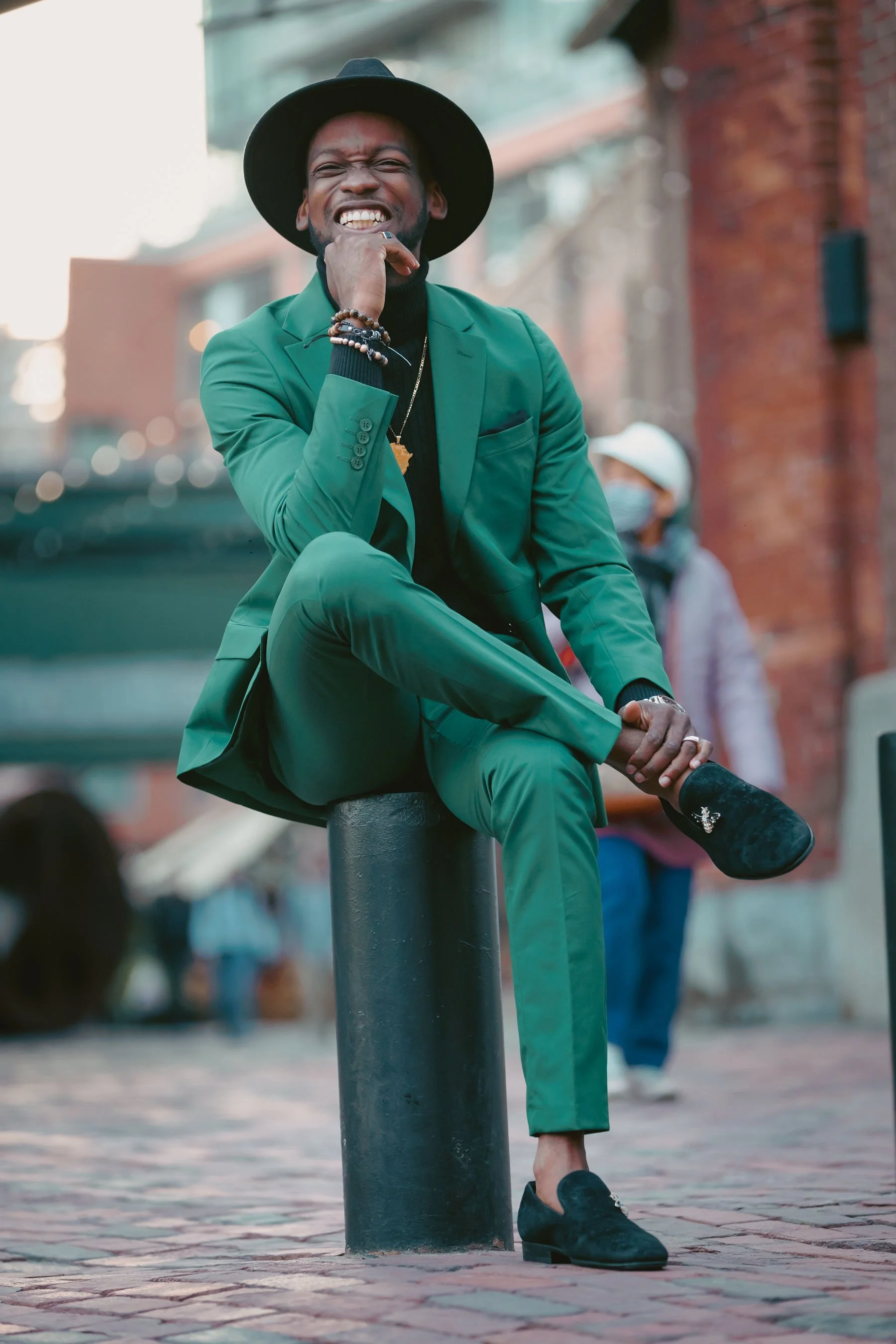 A man in a green suit and black hat sitting on a black bollard on a city street, smiling with his hand near his face, wearing bracelets and a gold chain. He embodies stylish, approachable financial coaching.