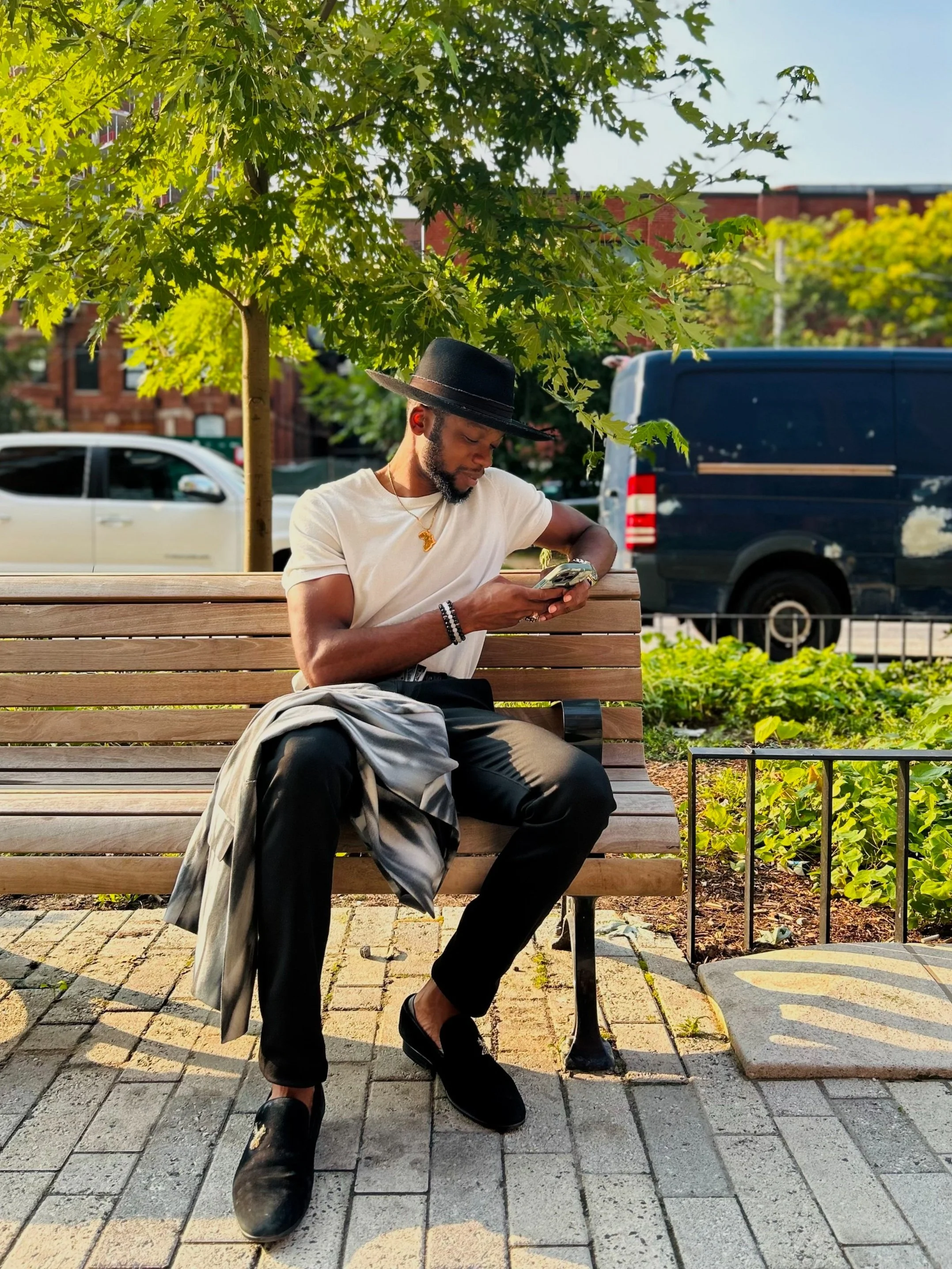 A man wearing a black hat and white t-shirt sits on a wooden park bench, looking at his phone with a jacket on his lap in an urban park — a modern financial coach staying connected on the go.