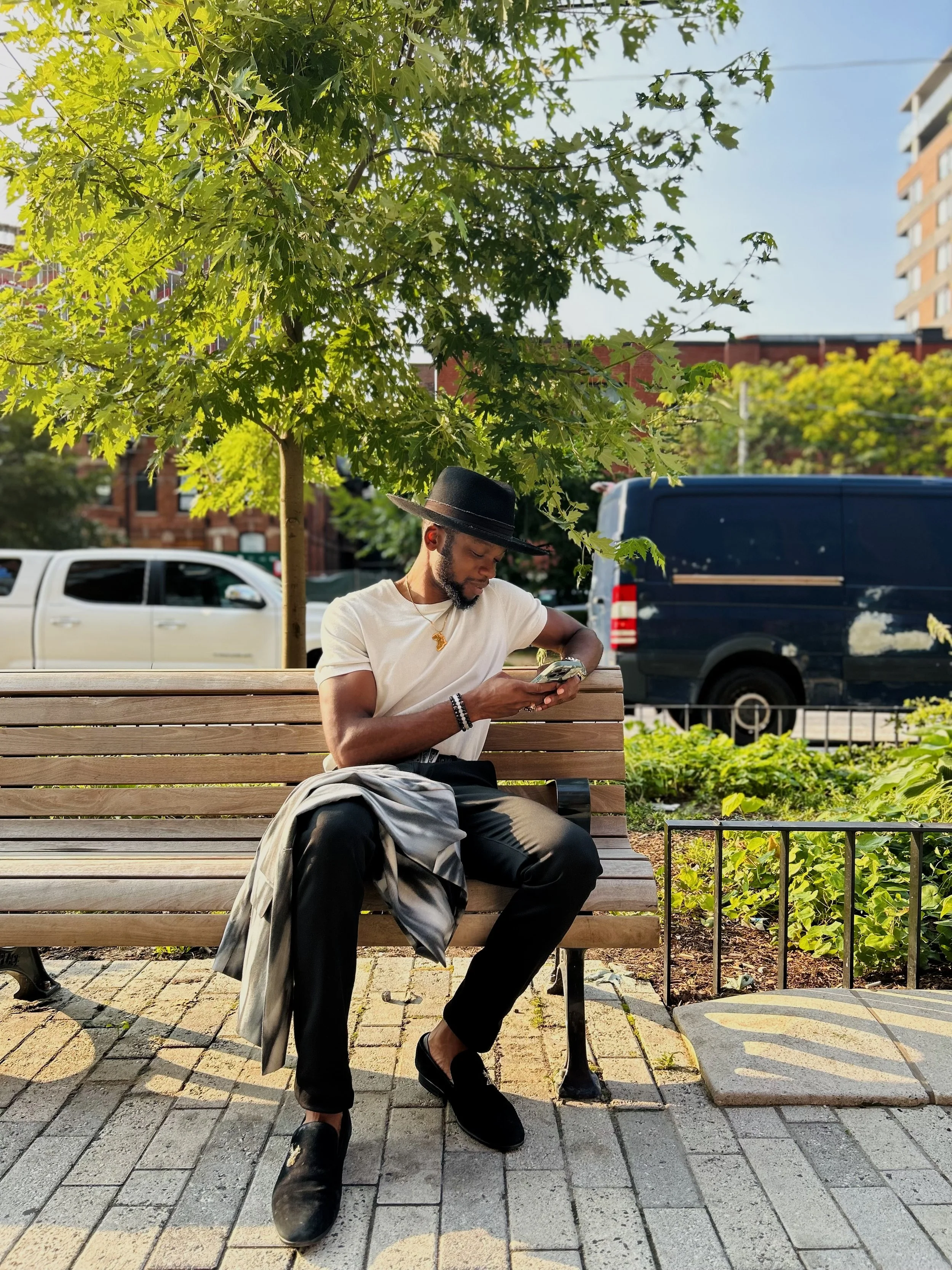 A man wearing a black hat and white t-shirt sits on a wooden park bench, looking at his phone with a jacket on his lap in an urban park — a modern financial coach staying connected on the go.