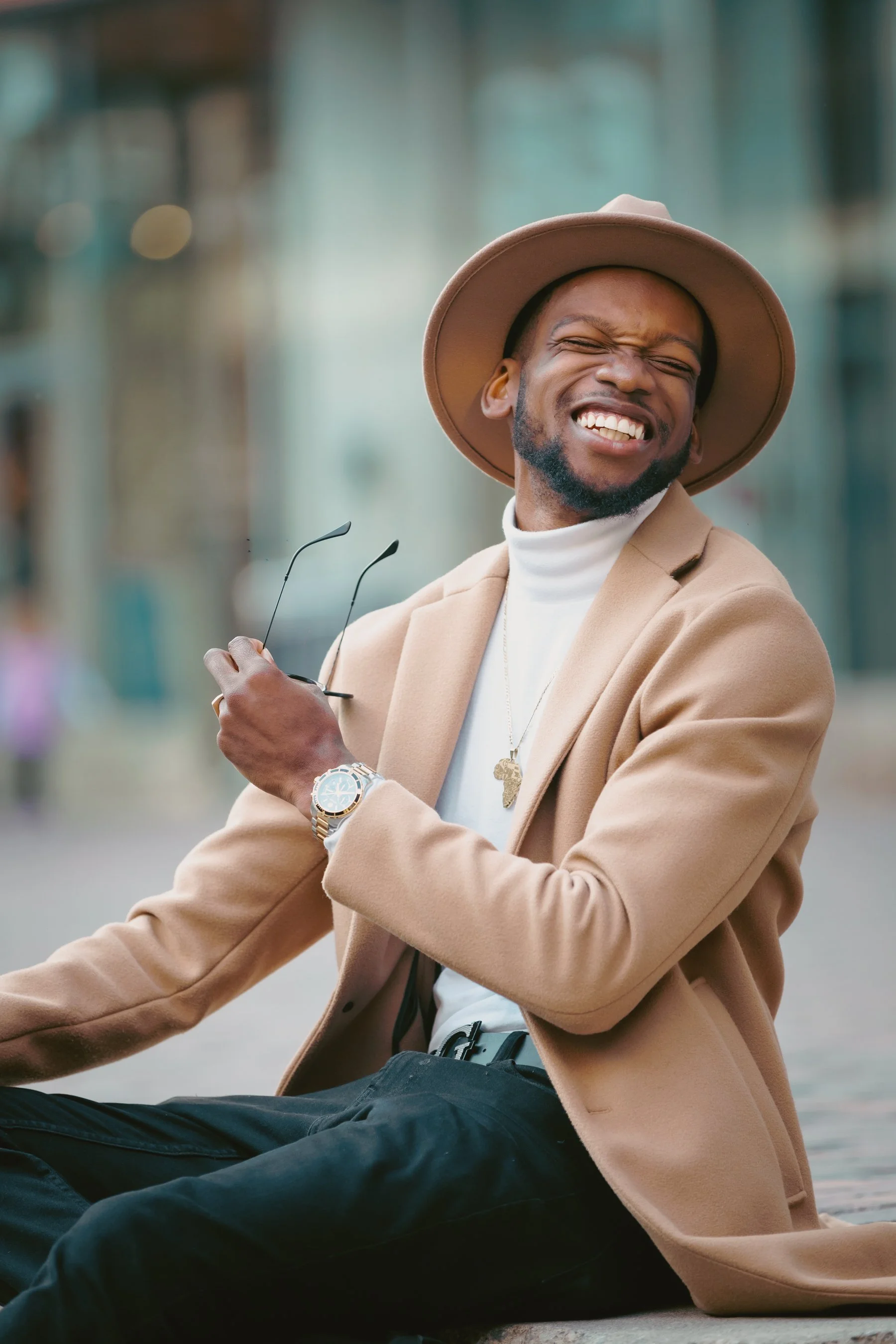 A young man in a tan coat and wide-brimmed hat sits outdoors with a bright smile, eyes closed, and sunglasses in hand — representing friendly, confident financial coaching with personality.