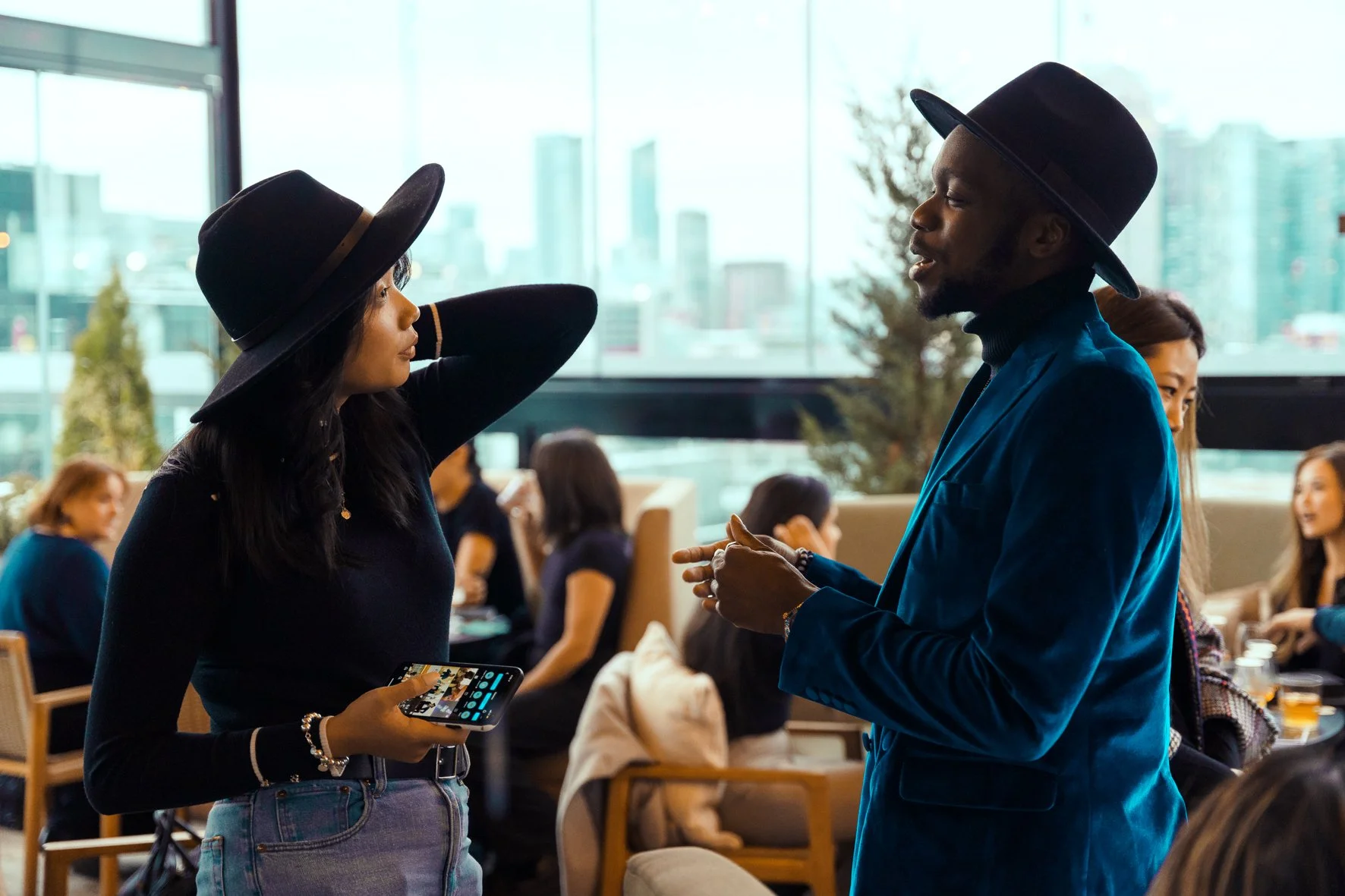 Two people talking in a bright office with large windows and city views. The woman adjusts her hat and holds a phone, while the man gestures—showing collaborative financial consulting and client support.