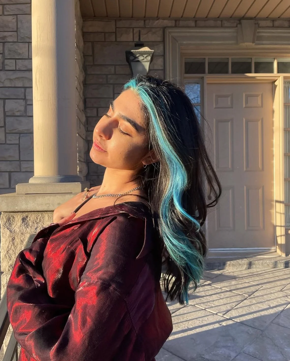 A woman with black and turquoise hair, wearing a maroon jacket, standing on a porch with eyes closed and face tilted toward sunlight — a peaceful, satisfied client moment.