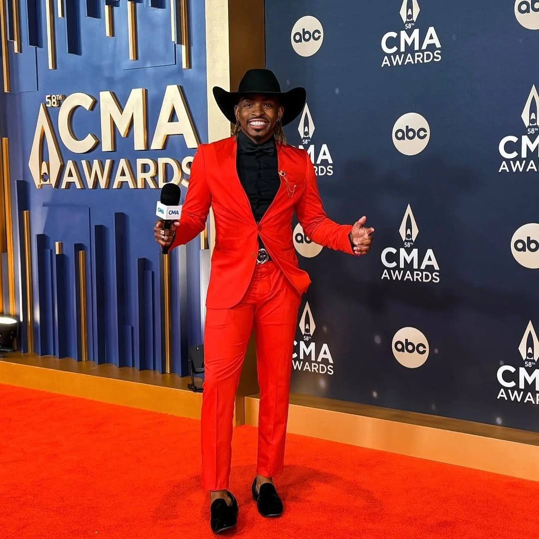 A man dressed in a bright red suit, black shirt, black hat, and black shoes, smiling and holding a microphone on the red carpet at the CMA Awards — showcasing a successful client.