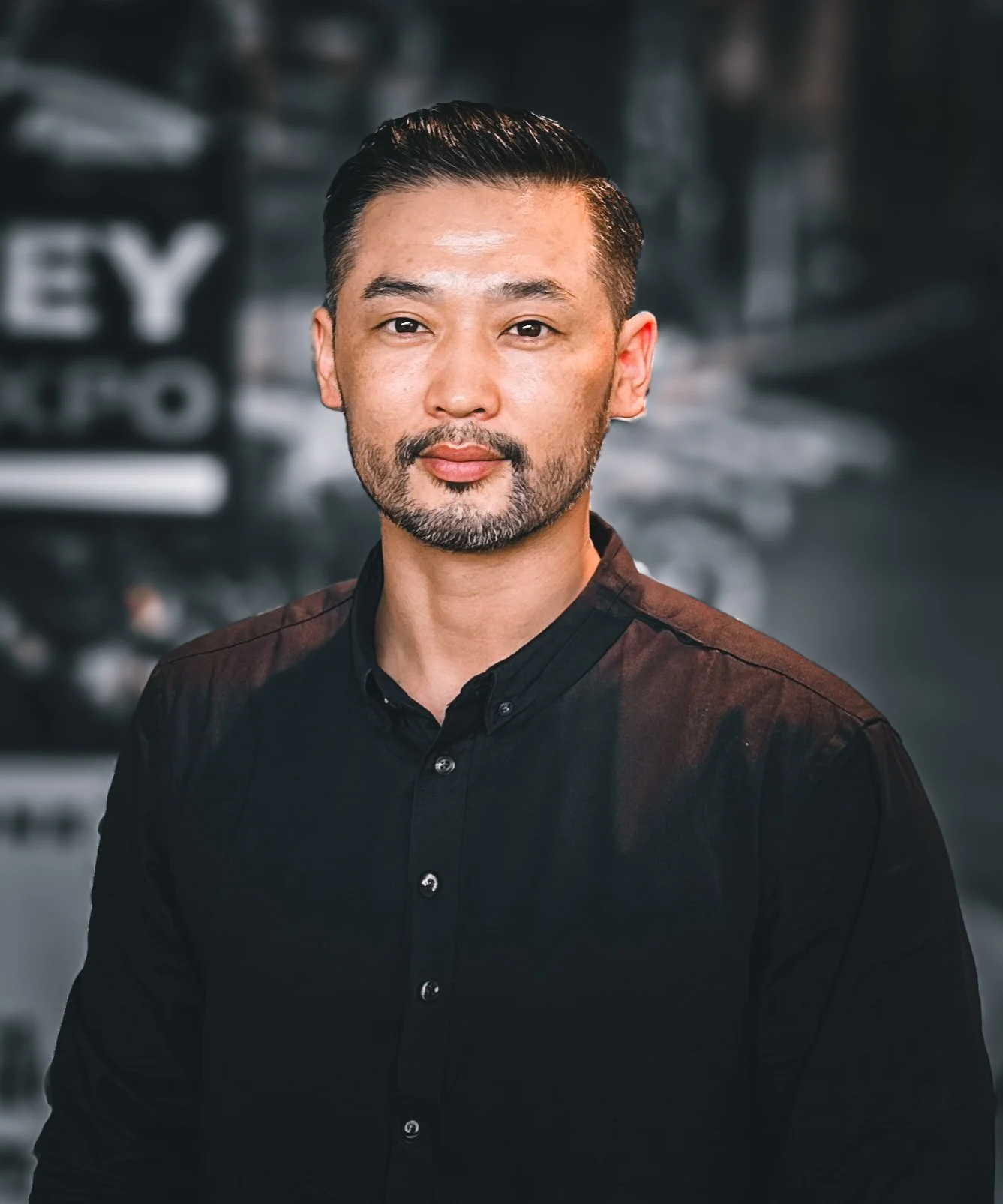 A man with short dark hair, a beard, and mustache, wearing a light blue and white striped polo shirt, standing indoors with a blurred background.