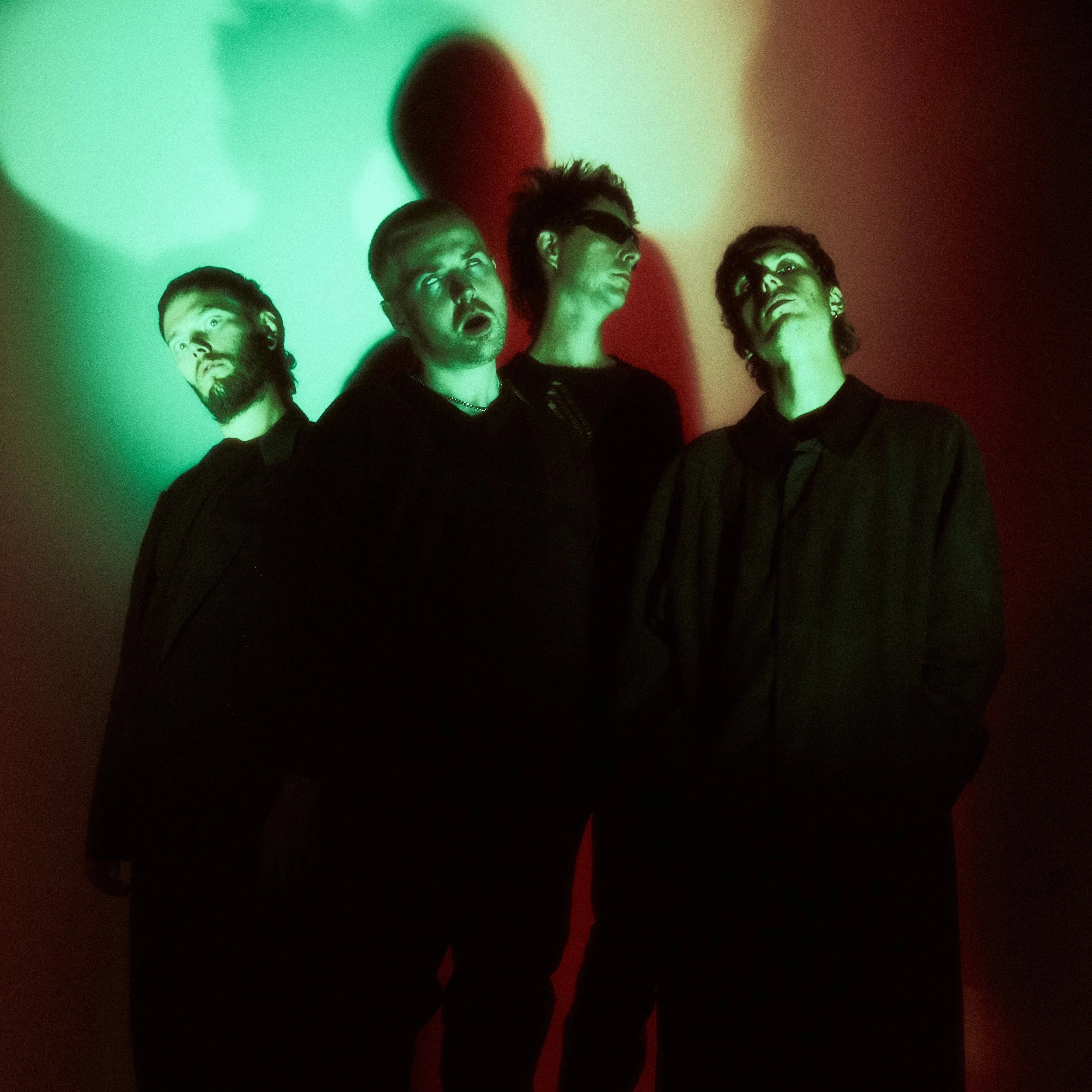 Four people with short hair and glasses stand in front of a wall lit with green, yellow, and red lights. Their shadows are cast on the wall.