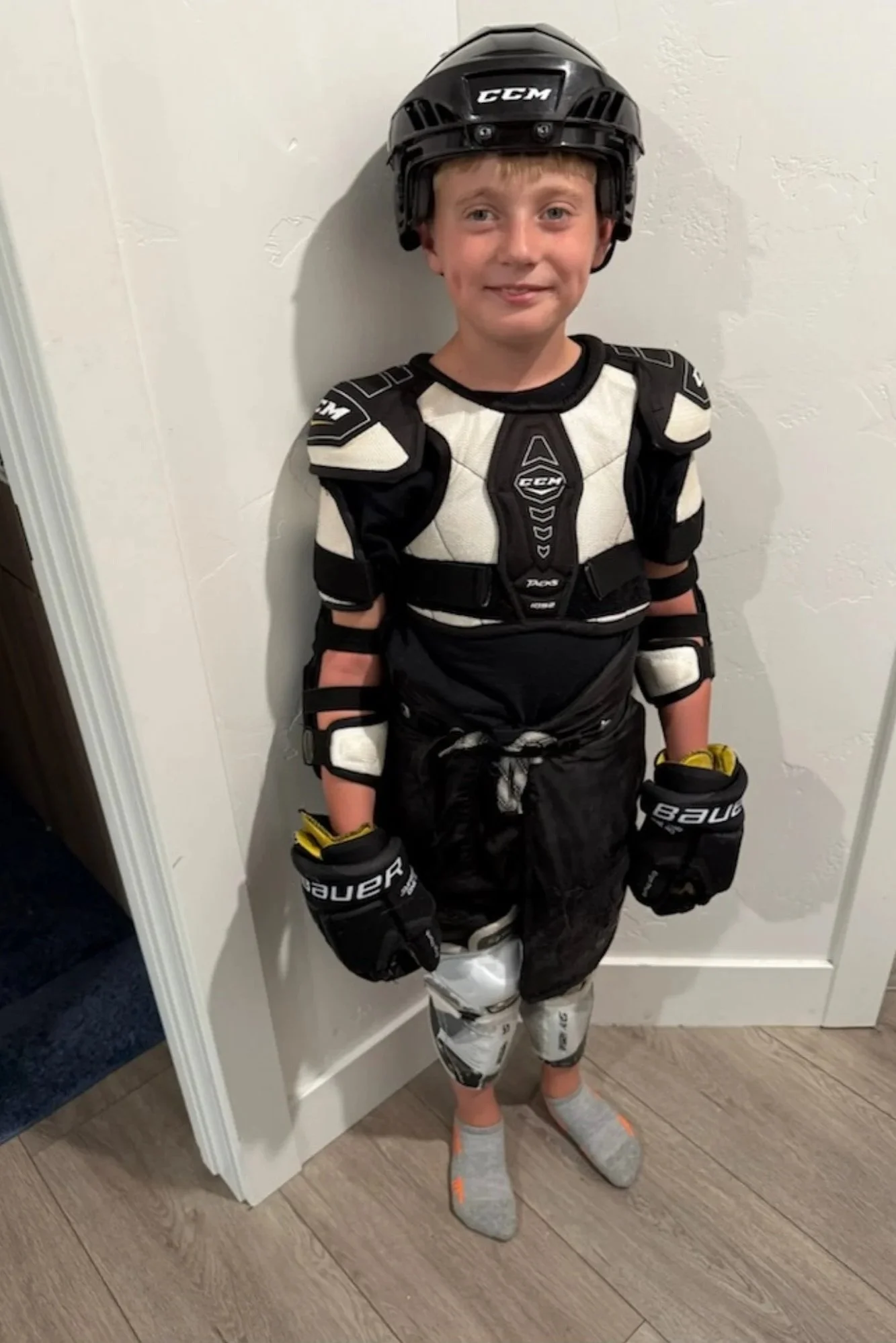 A young boy dressed in hockey gear, including a helmet, chest protector, gloves, hockey pants, and socks, standing against a white wall indoors.
