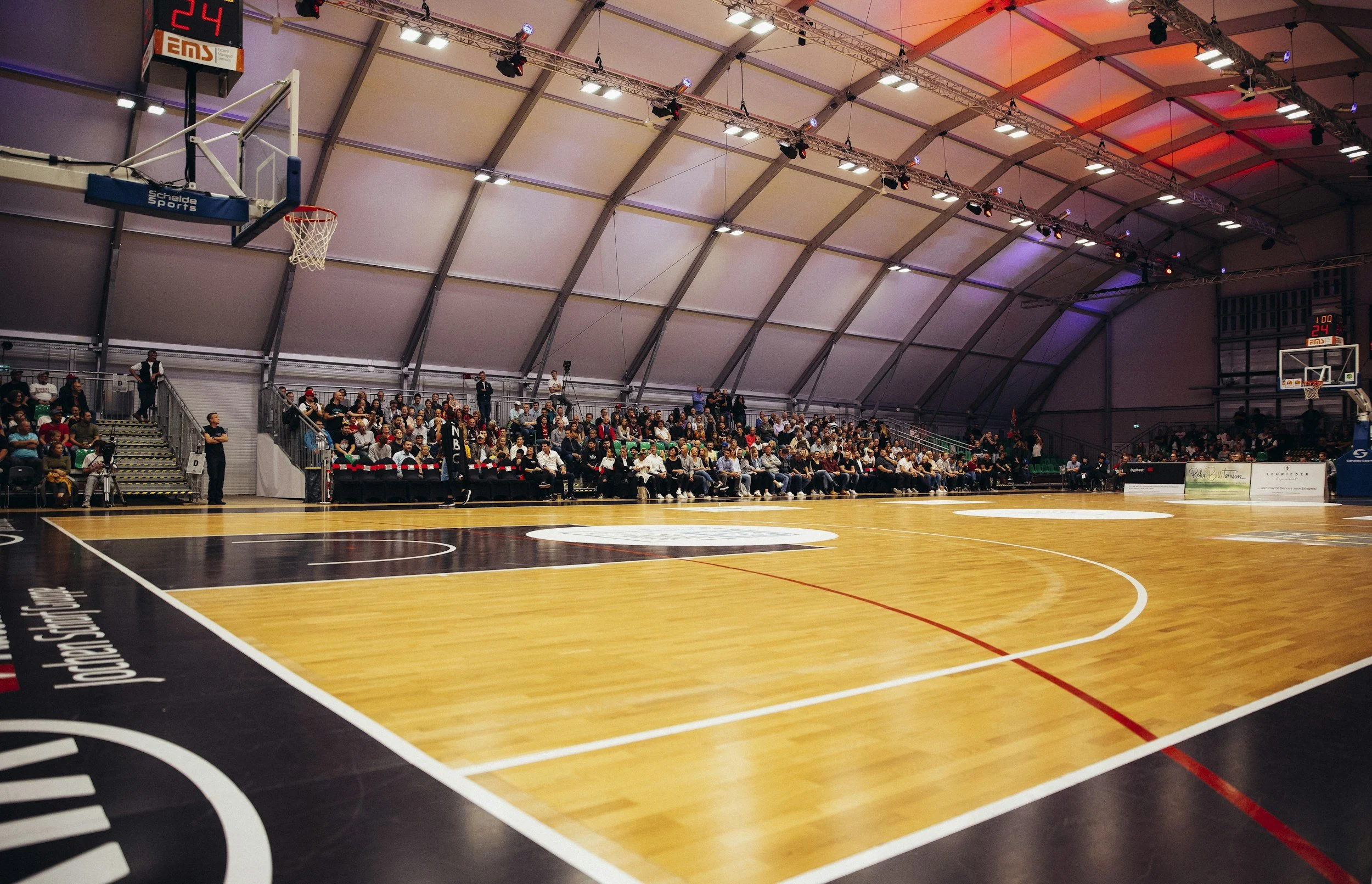 Indoor basketball court with spectators seated along the sidelines, scoreboard showing 24 seconds on the shot clock, and the court illuminated with colorful ceiling lights.