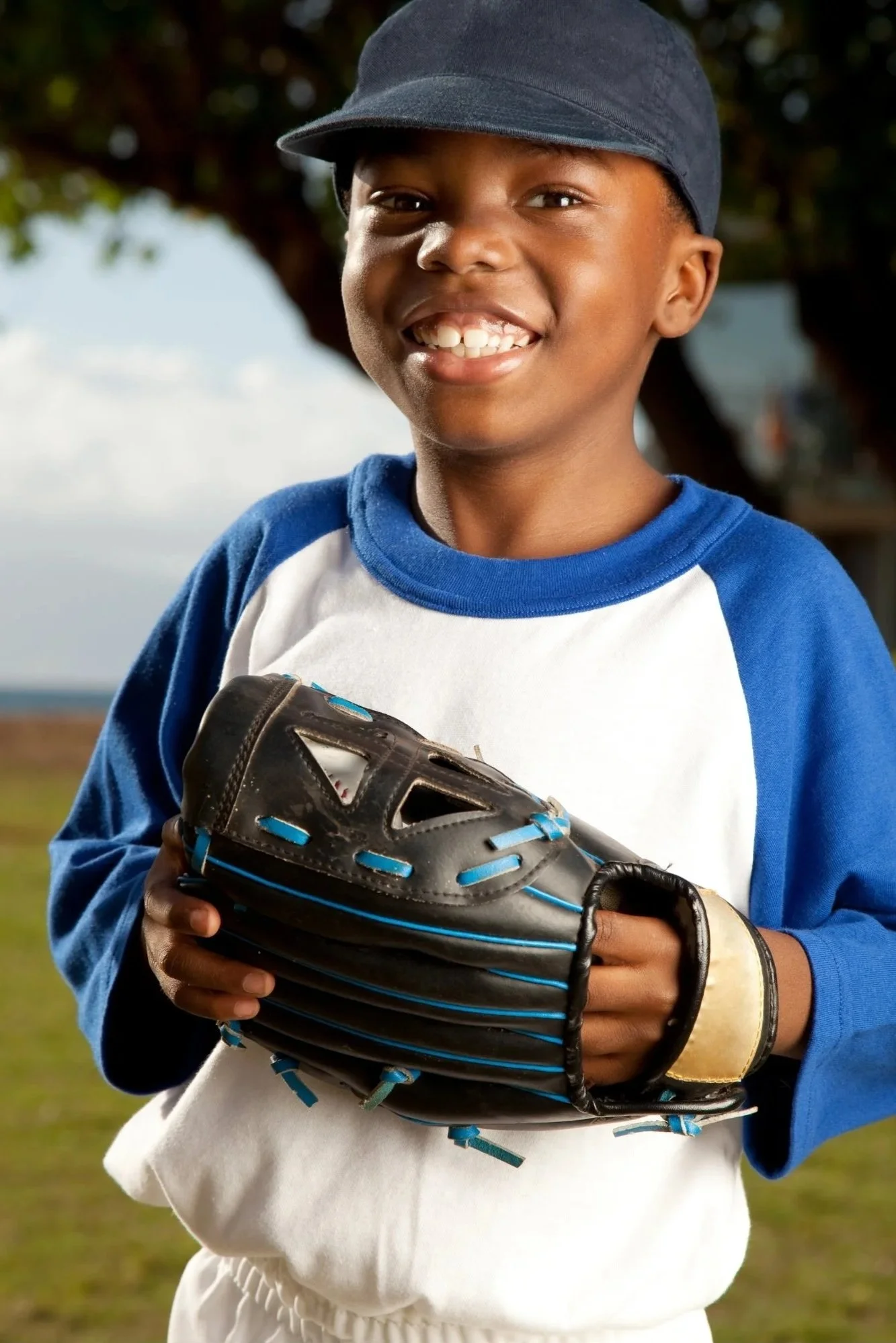 Young boy smiling outdoors holding a baseball glove, wearing a blue and white baseball uniform and a dark cap.
