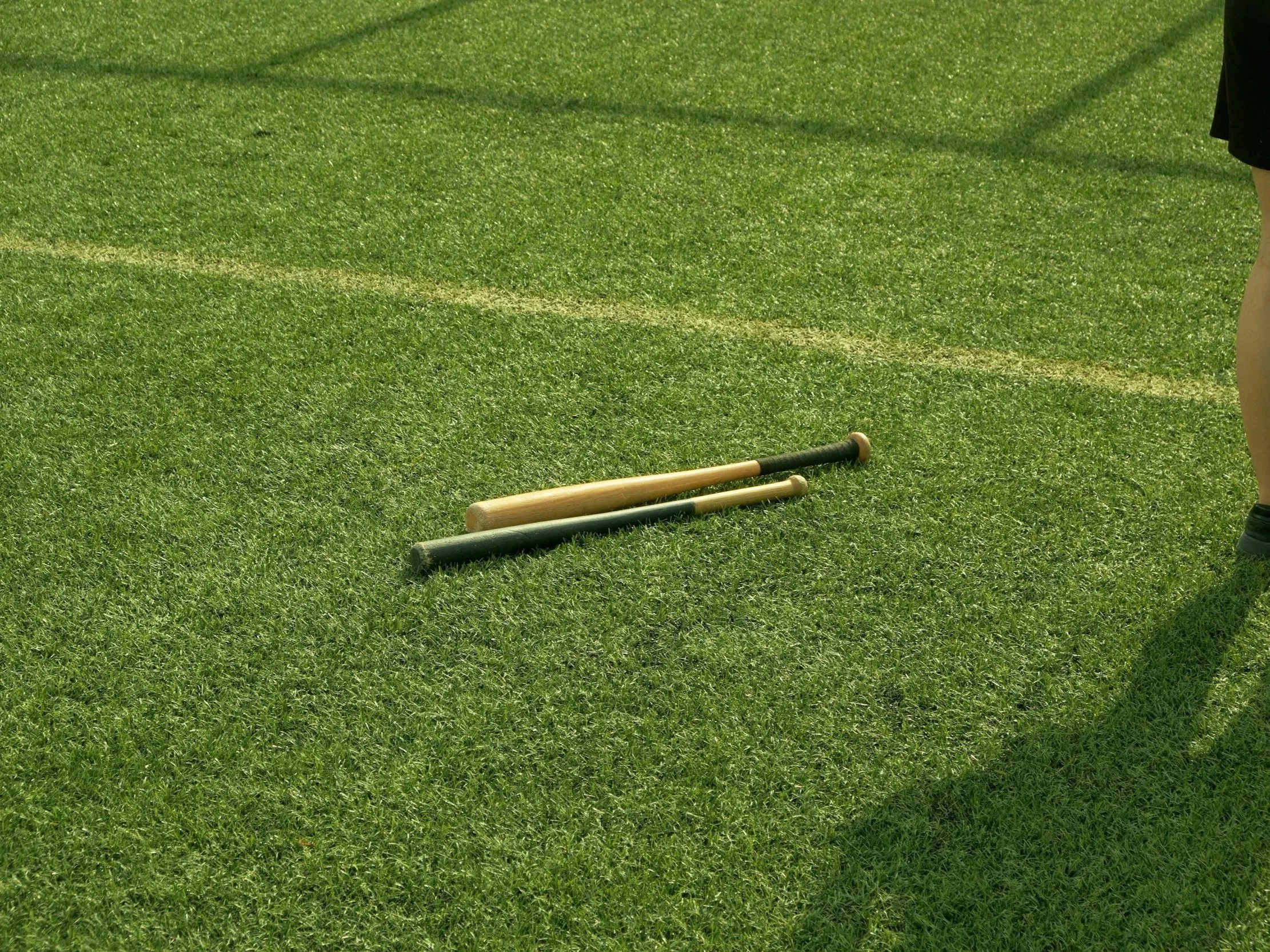 Two cricket bats lying on a grassy field, with a person standing nearby.