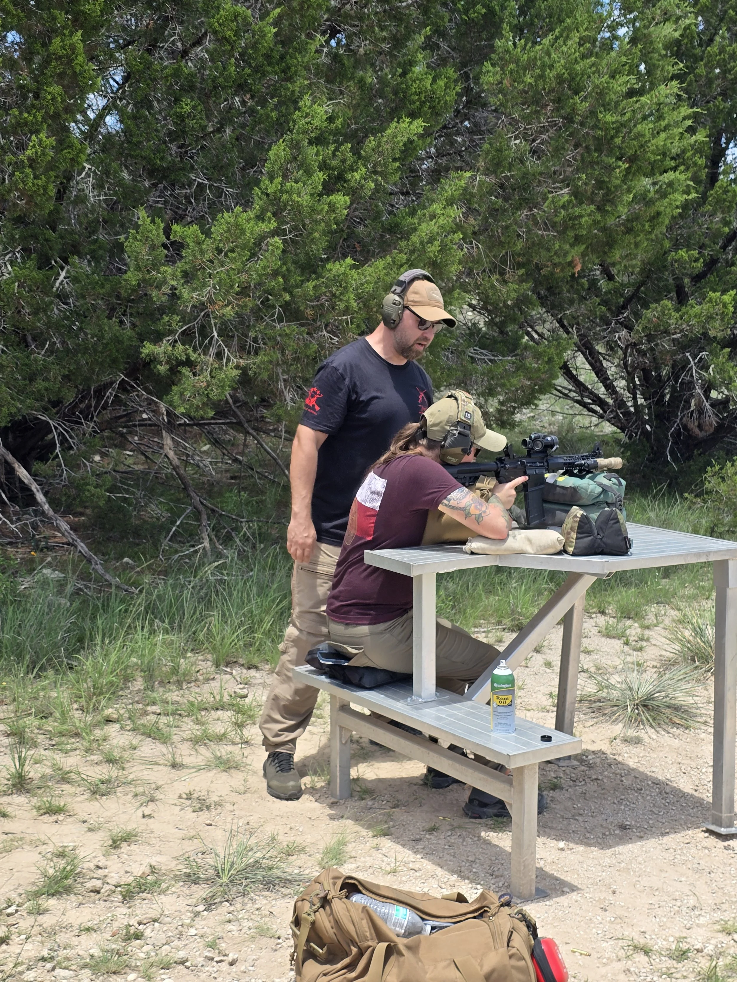 A man and a woman at an outdoor shooting range, with the woman aiming a scoped rifle on a shooting table and the man standing beside her, giving instructions or support.
