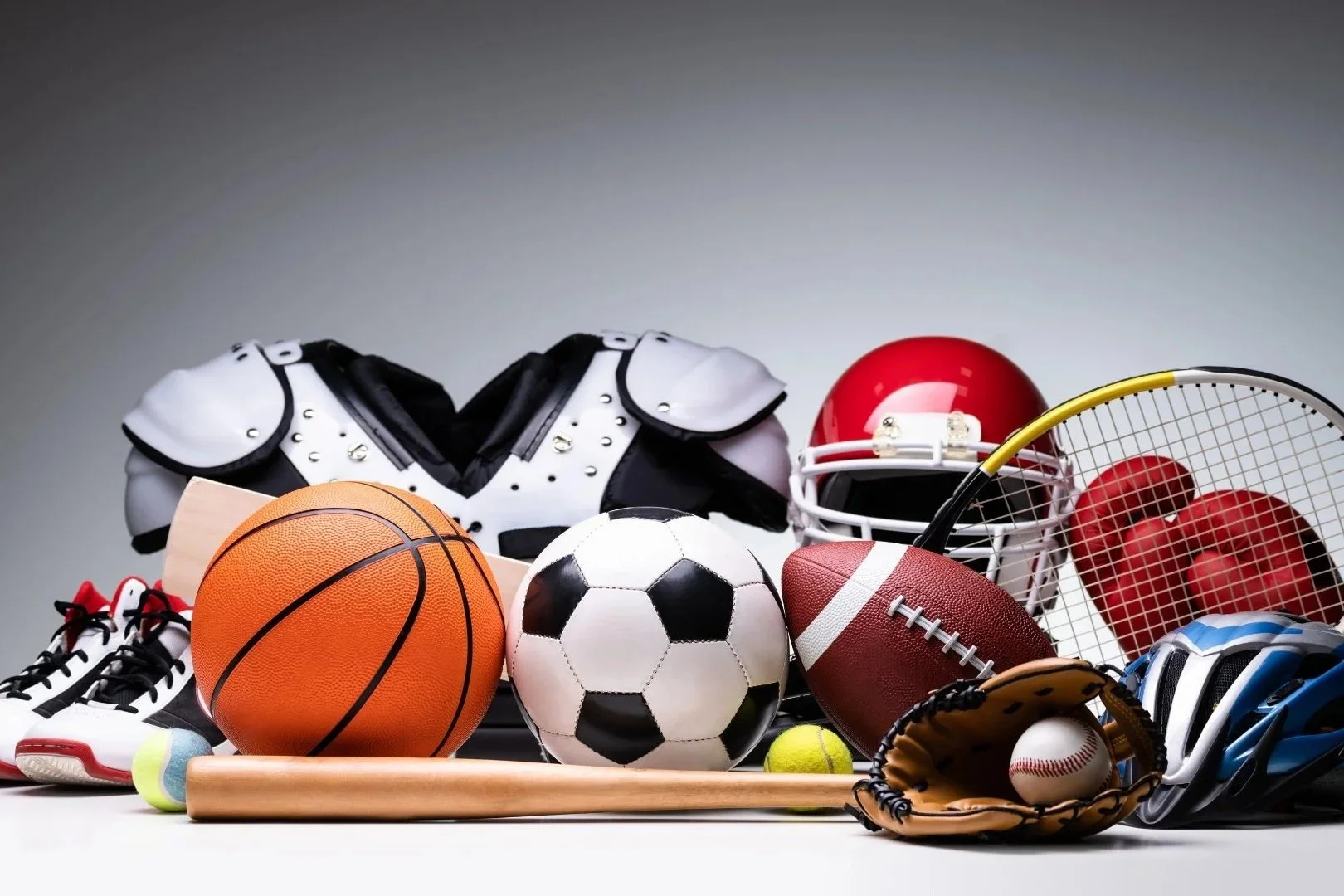 Collection of sports equipment including football helmet, basketball, soccer ball, football, tennis ball, baseball, baseball glove with ball, tennis racket with balls, cleats, catcher's gear, and various sports balls.