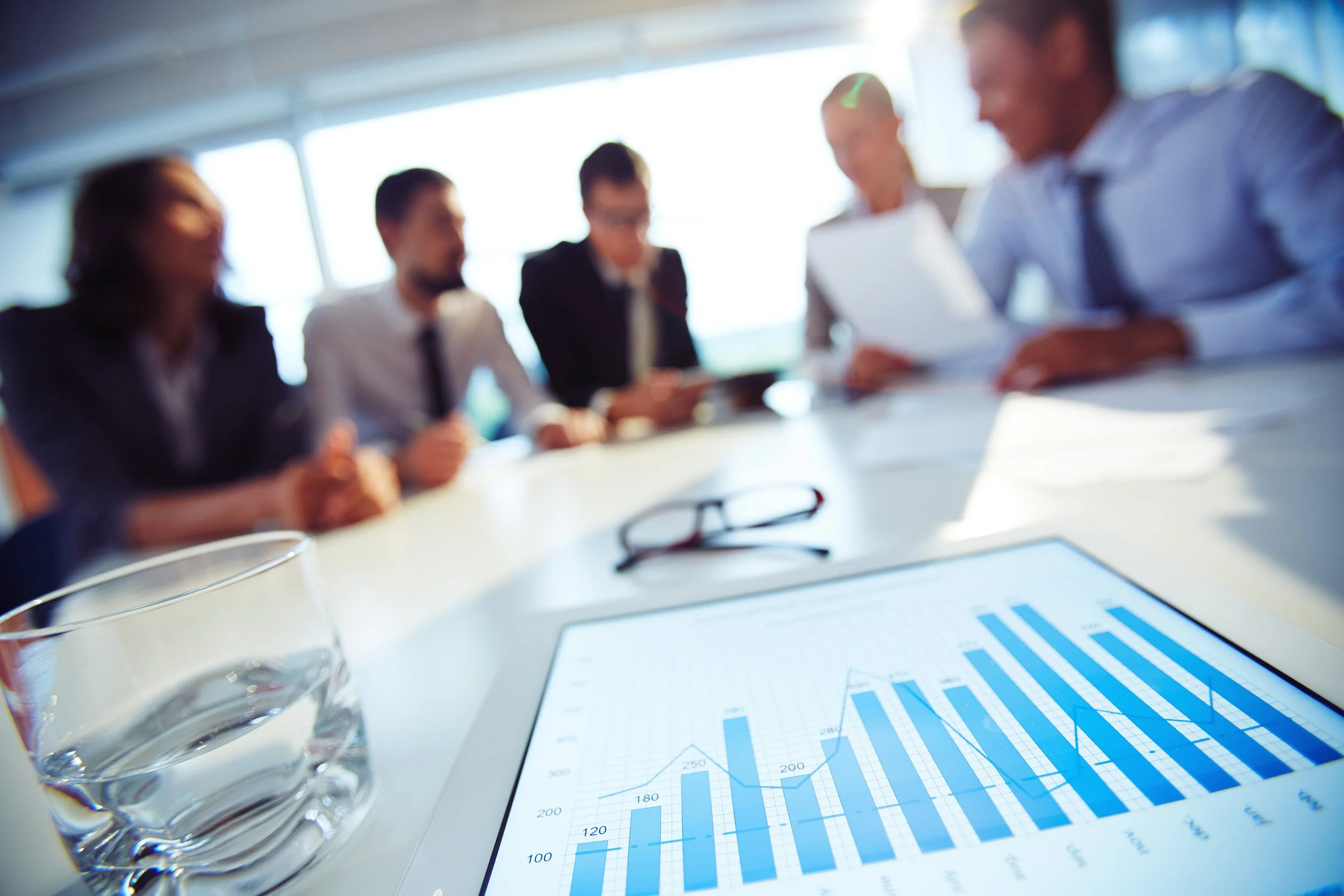 Un groupe de professionnels en réunion d'affaires. Sur la table, un écran affiche un graphique de barres bleues indiquant des statistiques croissantes. À côté, un verre d'eau et une paire de lunettes. La pièce est bien éclairée par de grandes fenêtres.