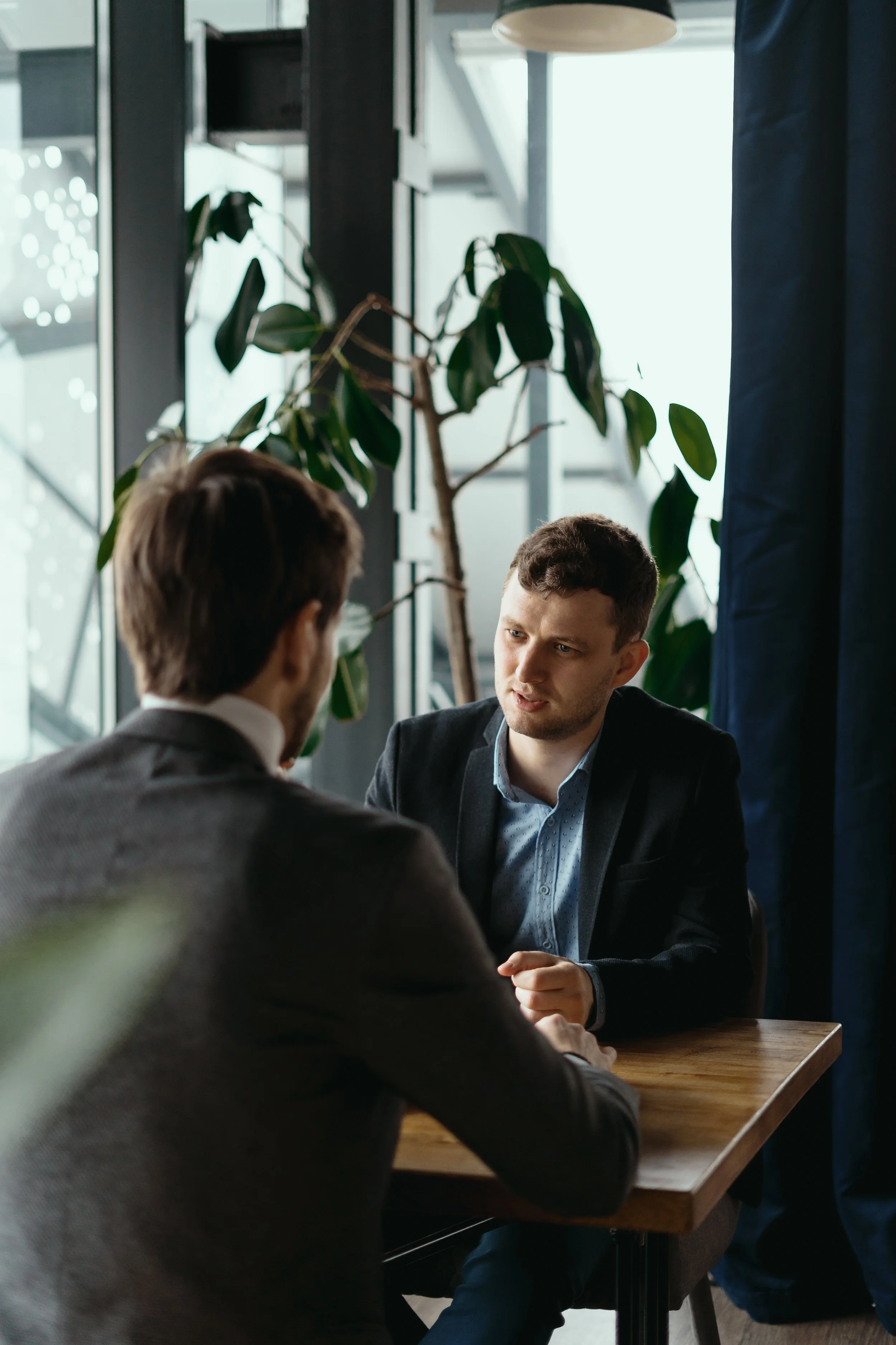 Deux hommes en costume émire discutent dans un café ou un espace de travail, avec une grande plante et une fenêtre en arrière-plan.