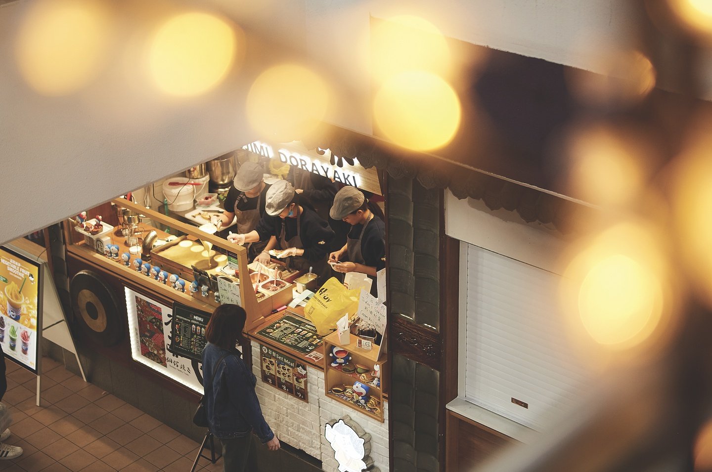 Aerial view of a Japanese street food vendor with staff preparing dorayaki, a customer standing nearby, and decorative lights in the foreground.