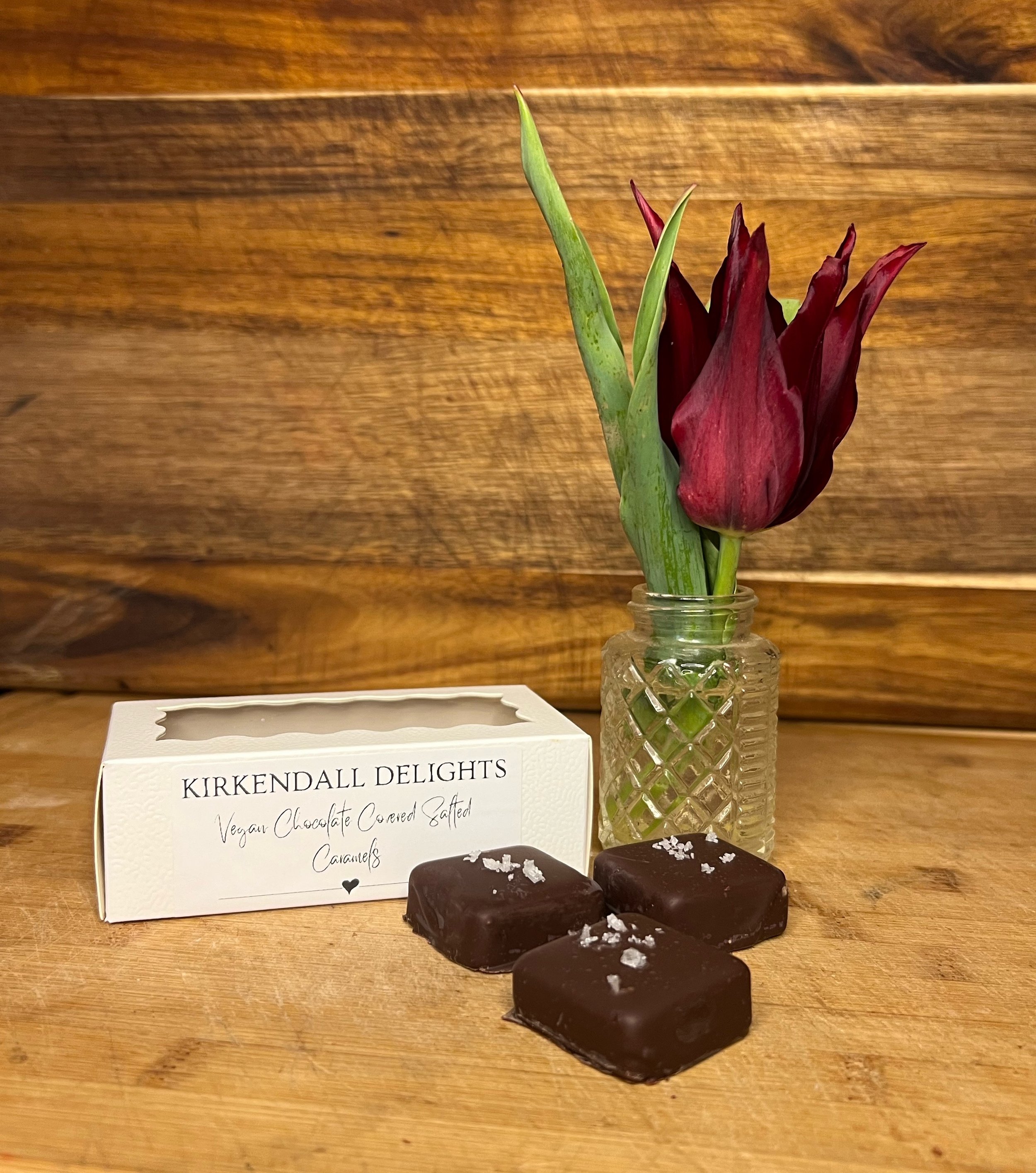 Box of Kirkendall Delights vegan chocolate-covered sea salt caramels beside a red tulip in a glass vase, displayed on a wooden counter