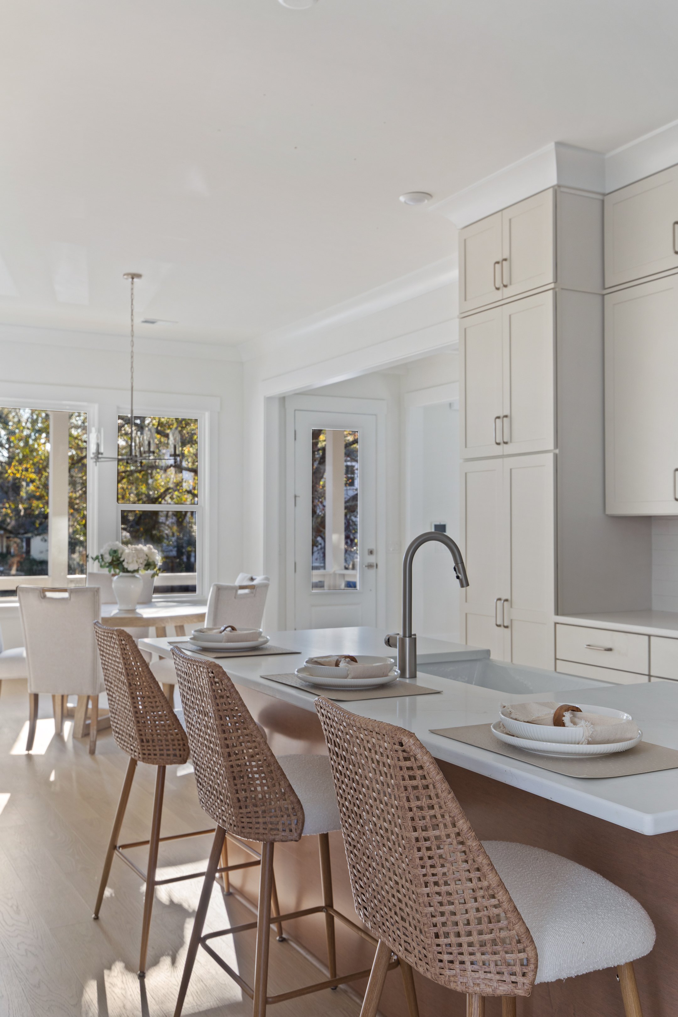Bright and airy kitchen with a large island, woven bar stools, and a dining area with a round table and white chairs, large windows showing trees outside.