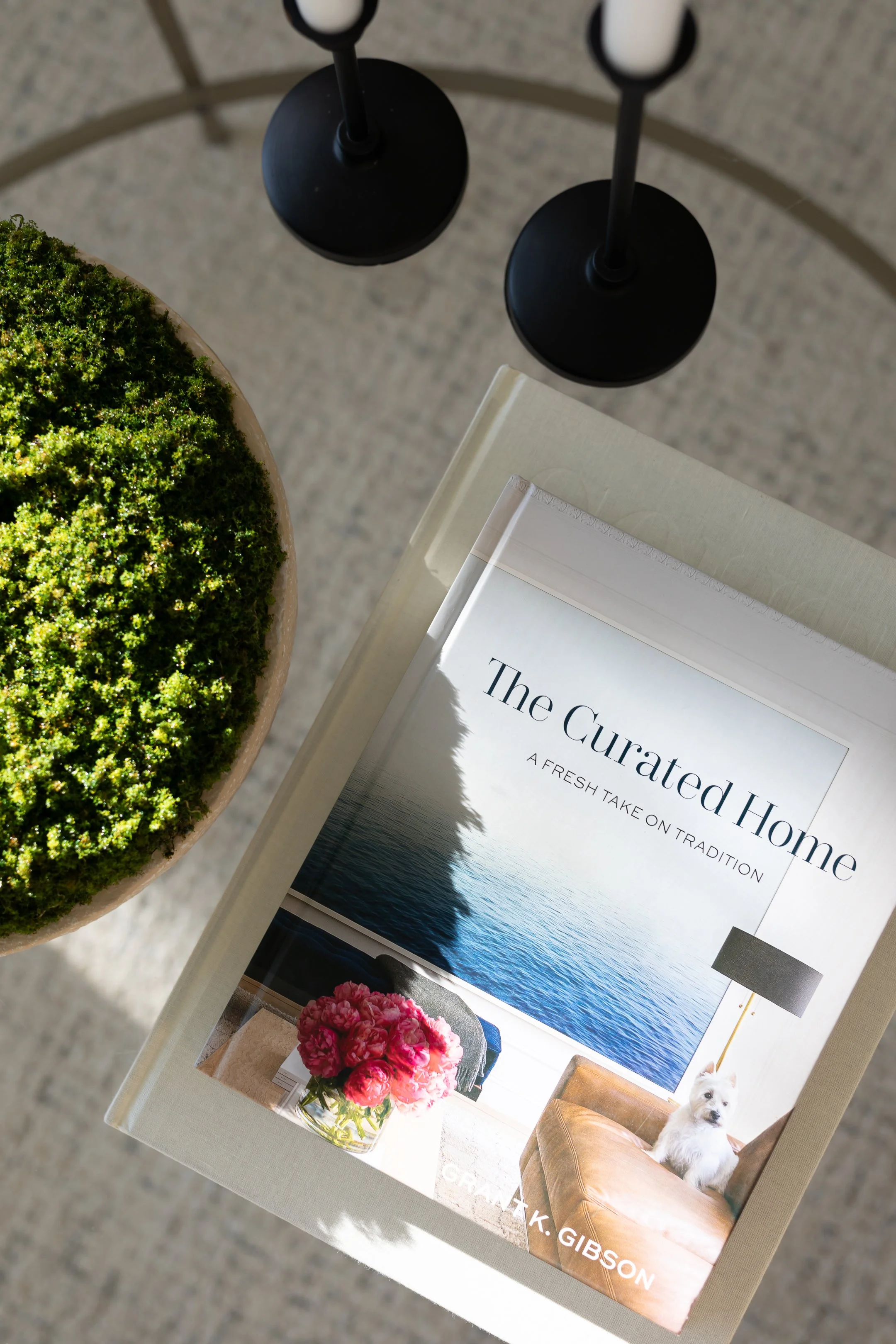 A book titled "The Curated Home" with the subtitle "A Fresh Take on Tradition," placed on a light-colored table next to a pot of green moss and a black floor lamp with two shades. The cover features a photo of a vase of pink flowers, a leather chair, and a small dog.