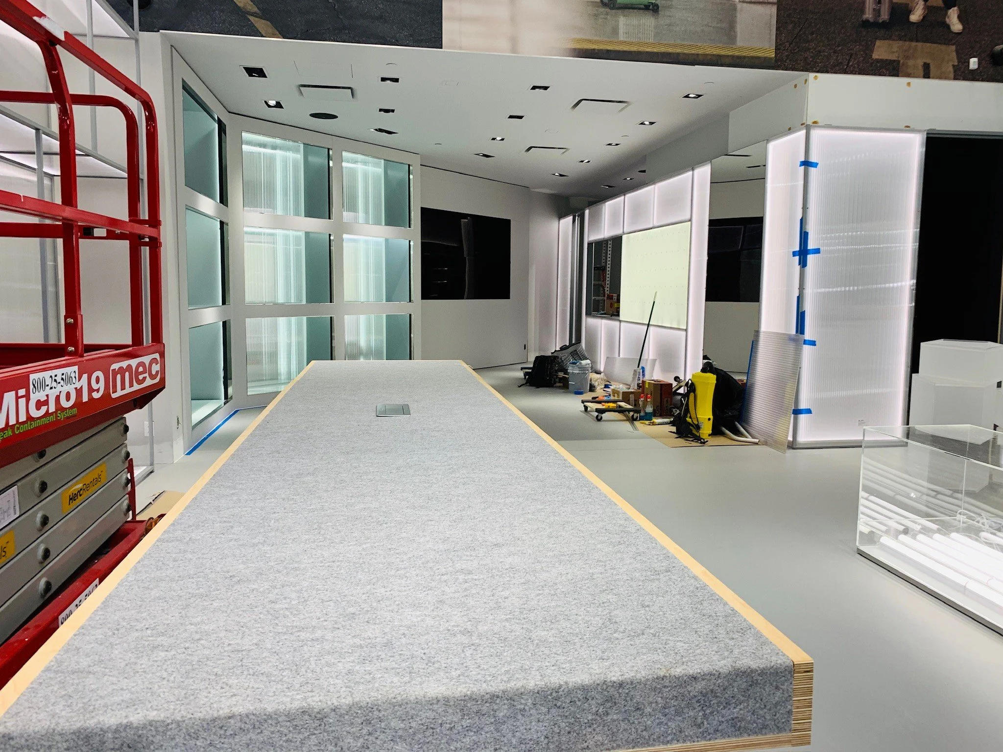 Interior of a construction site showing a partially completed room with a large white table in the foreground, various tools and equipment along the back wall, glass panels, and metallic frame structures.