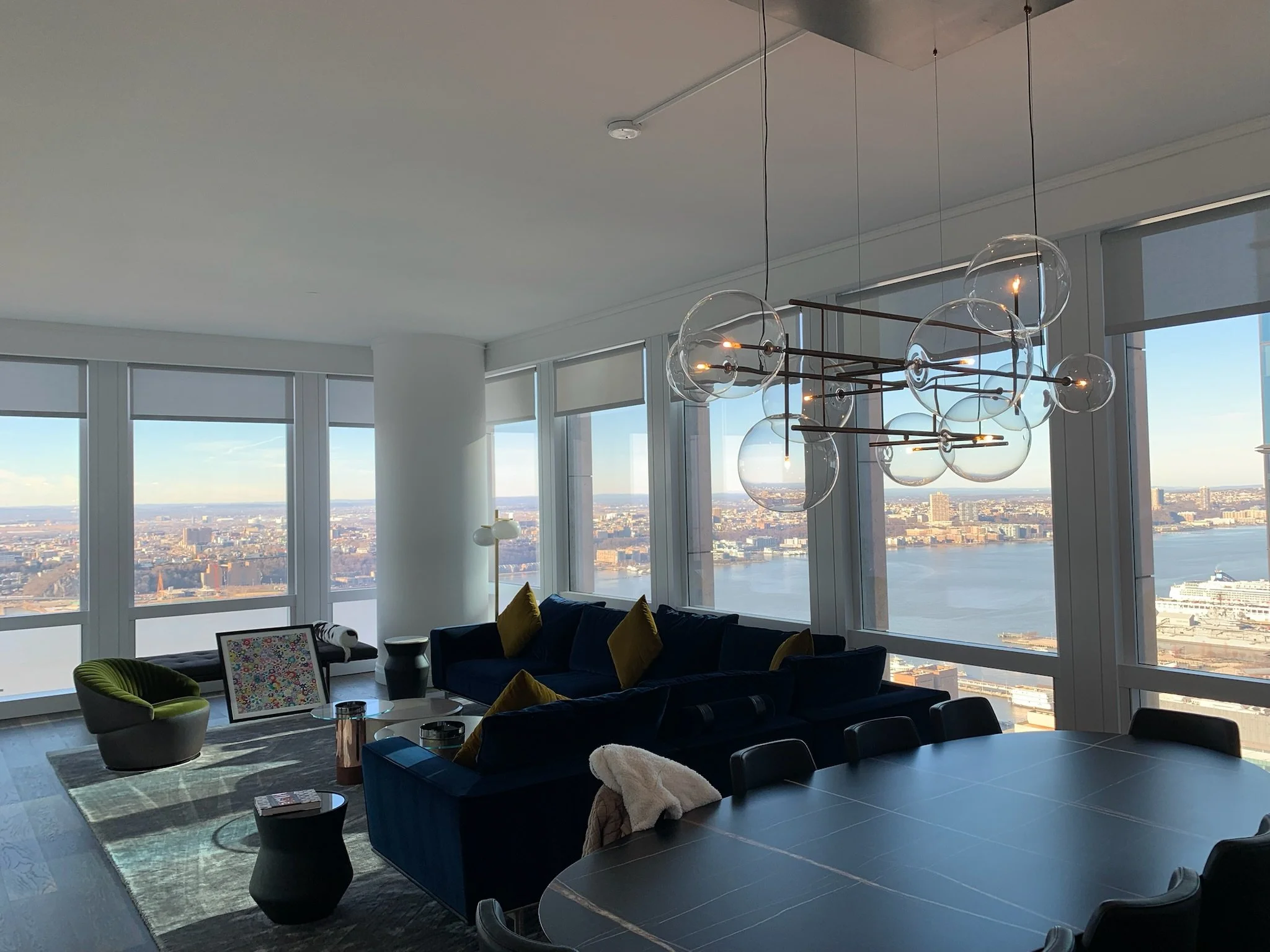 Modern high-rise living room with large floor-to-ceiling windows offering a city skyline and river view, featuring a dark blue sectional sofa with yellow cushions, a green accent chair, a round black side table, a coffee table, and a contemporary chandelier with multiple glass globe lights.