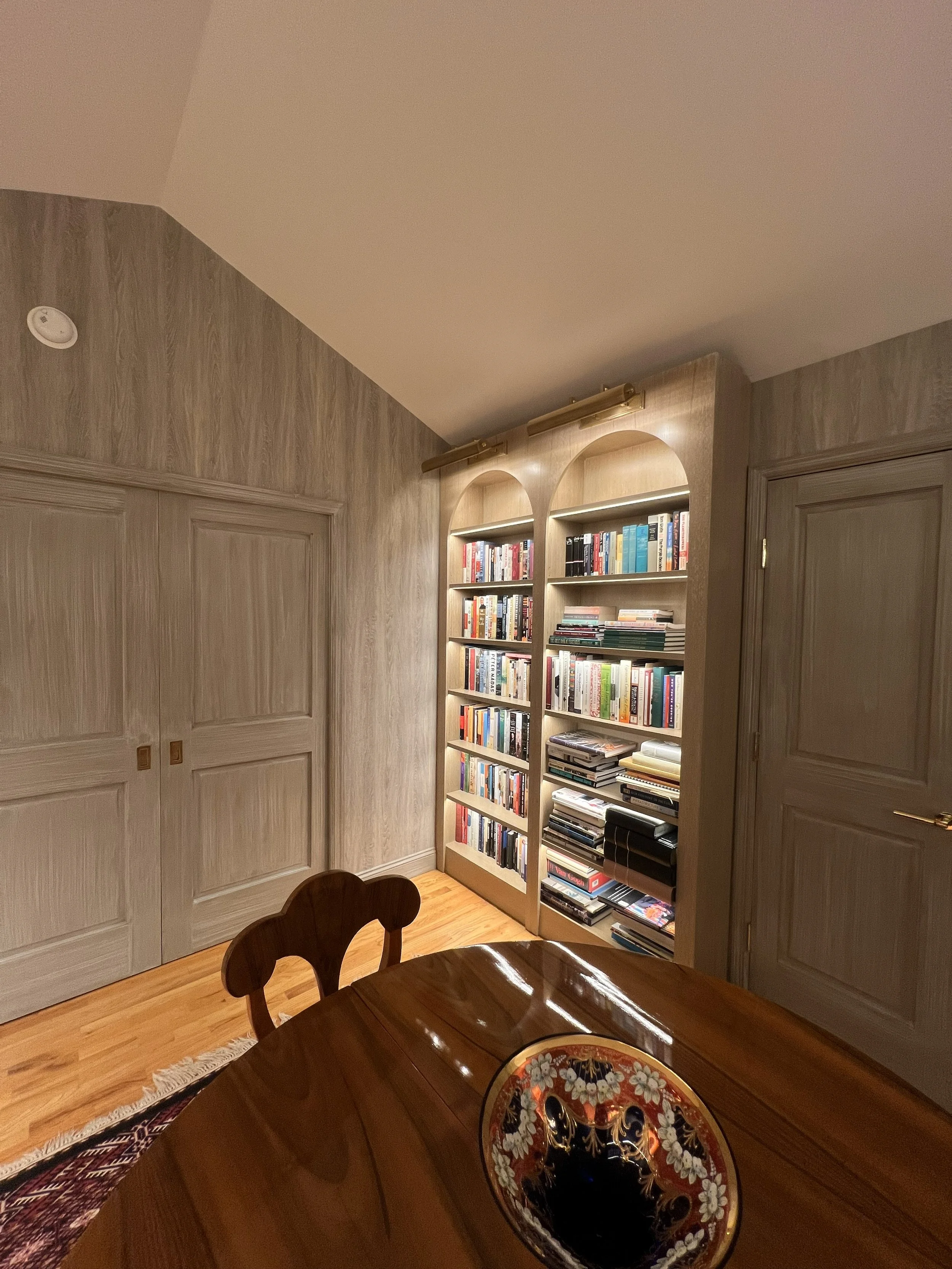 A custom wooden bookshelf filled with books located against a beige wall in a room with wooden flooring. Part of a wooden dining table and chair are visible in the foreground.