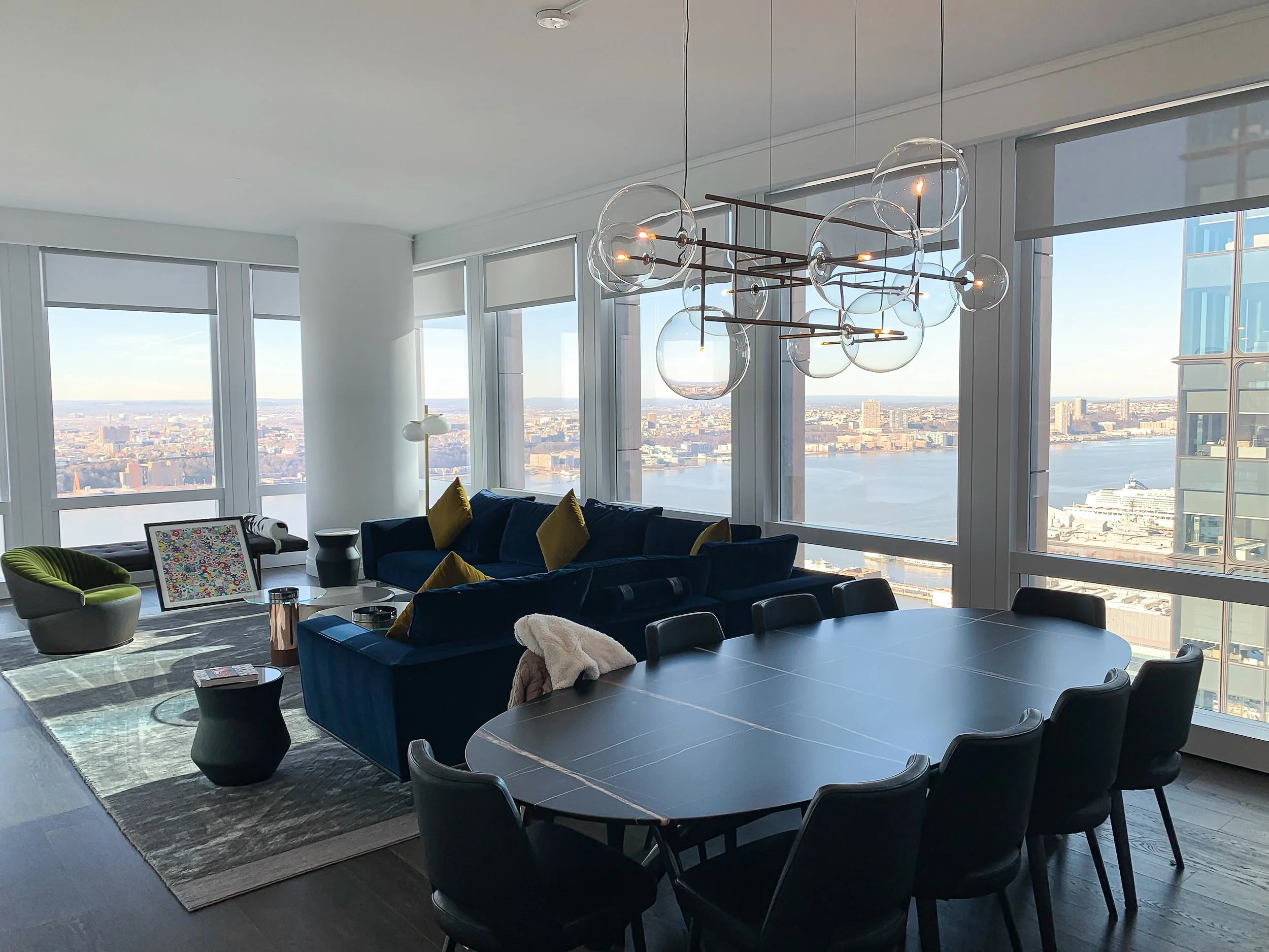Modern living room with floor-to-ceiling windows offering a city and river view, featuring a blue sectional sofa with yellow pillows, a round black dining table with eight black chairs, a green accent chair, a abstract pattern armchair, and stylish h