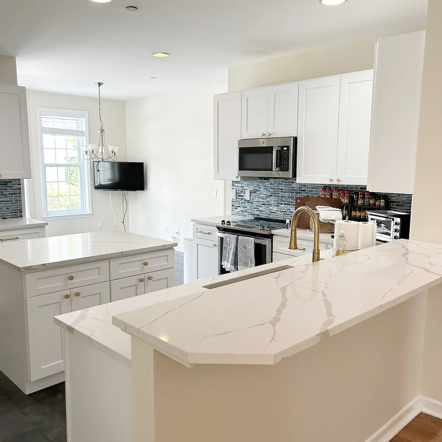 Modern white kitchen with marble countertops, stainless steel appliances, black mosaic backsplash, window, and a wall-mounted flat-screen TV.