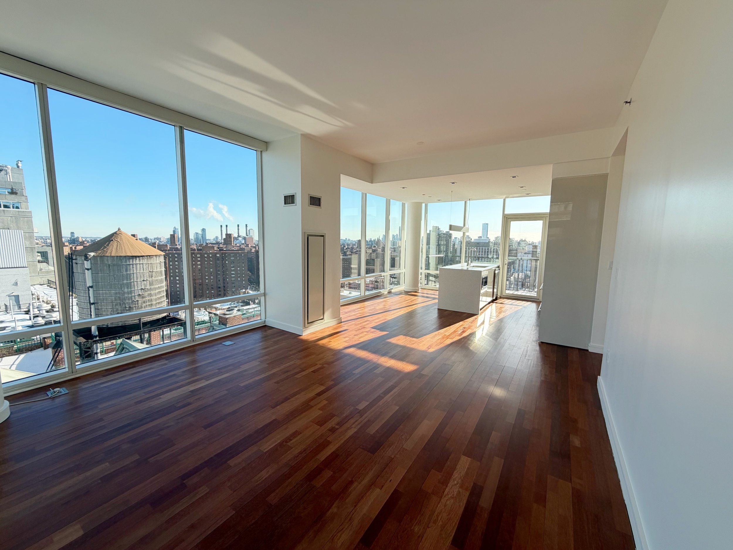 Empty modern apartment living room with large floor-to-ceiling windows showing city skyline and wooden floors.
