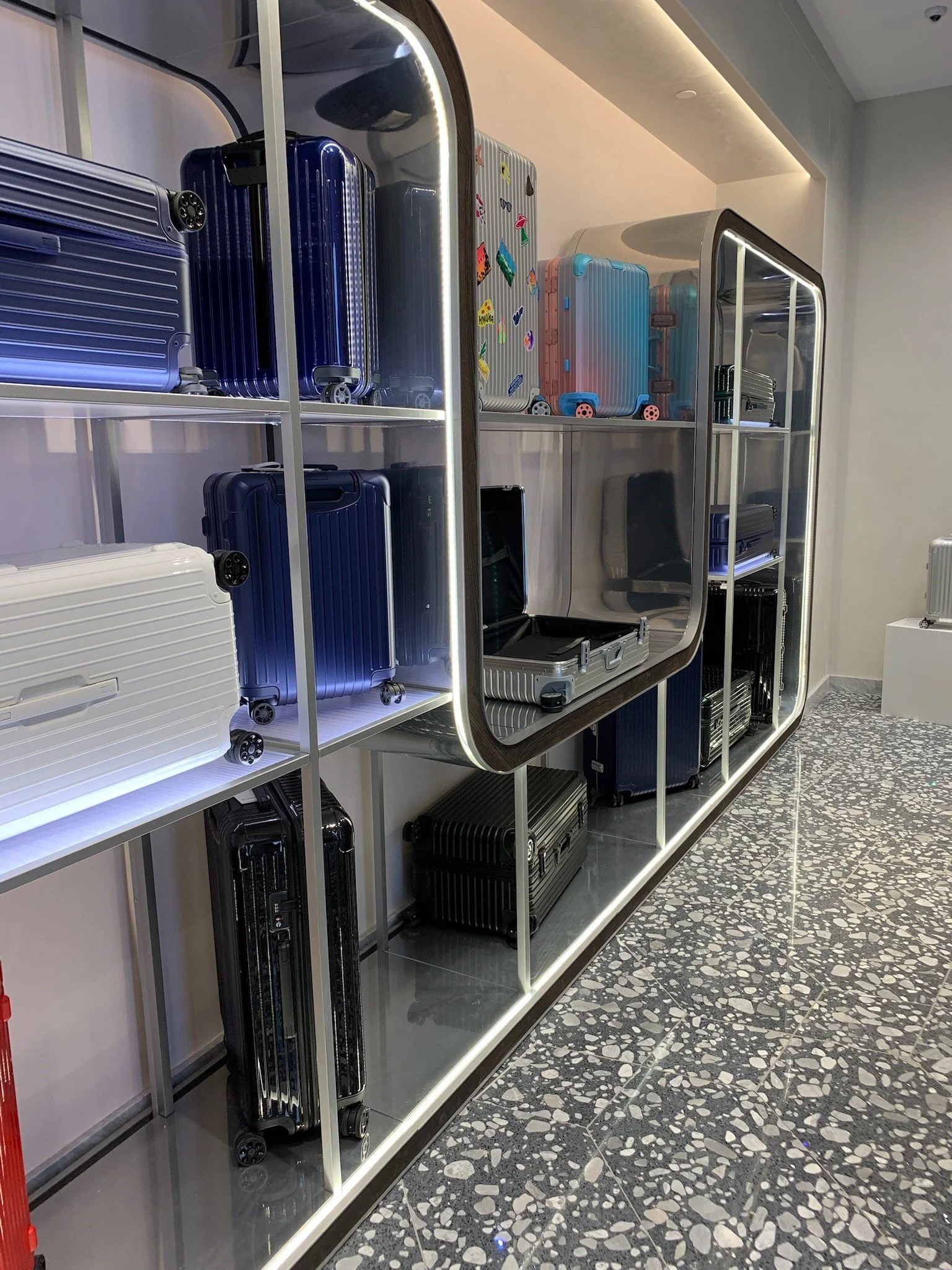 Display shelf with various suitcases in different colors and sizes, some with stickers, in a modern interior with terrazzo flooring.
