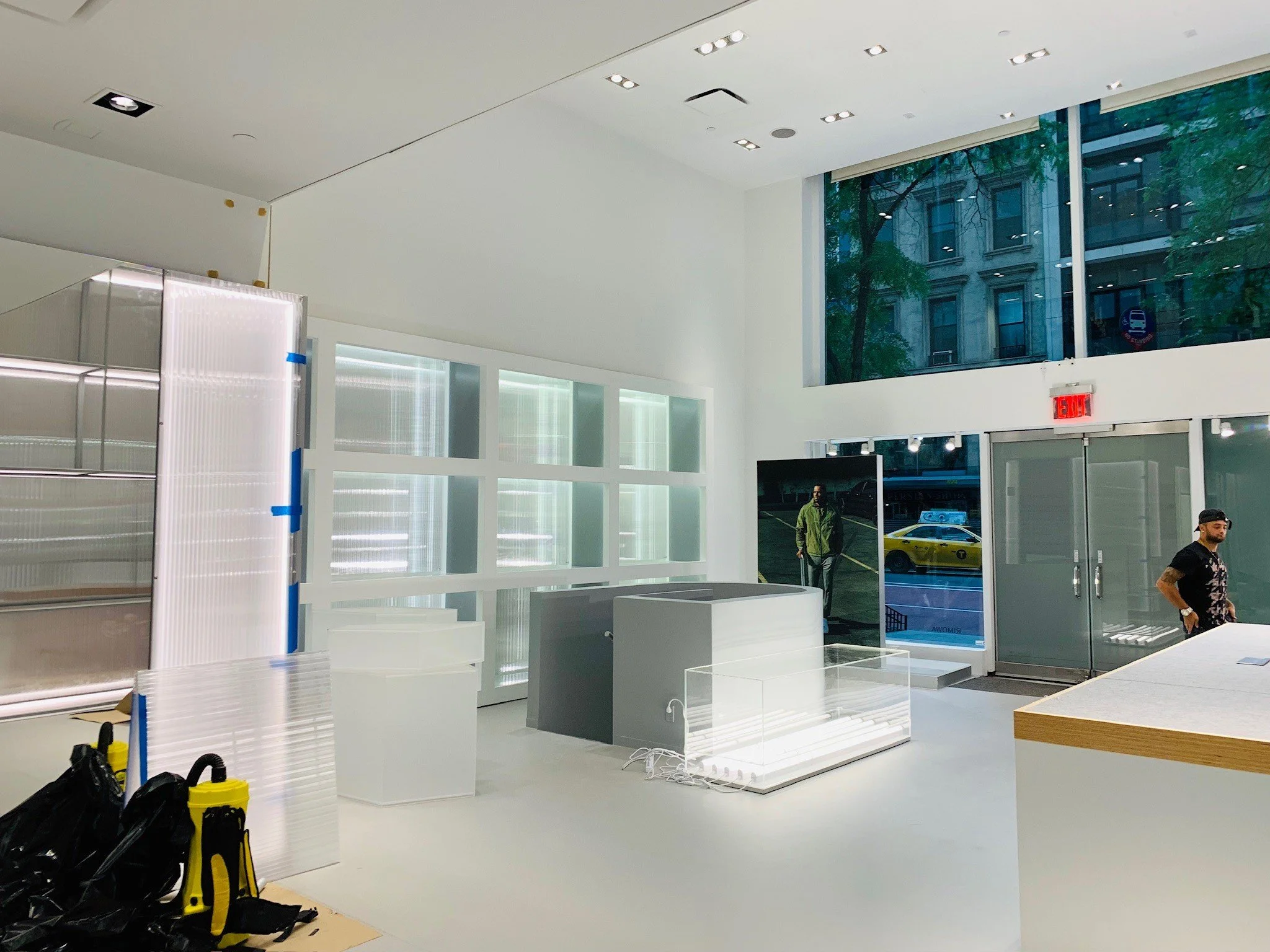 Empty retail or gallery space with white walls, large glass windows, and a doorway. Some manikins and a large empty display case visible. Man with a beard and black clothing stands near the glass doors, and another person is reflected in a digital sc