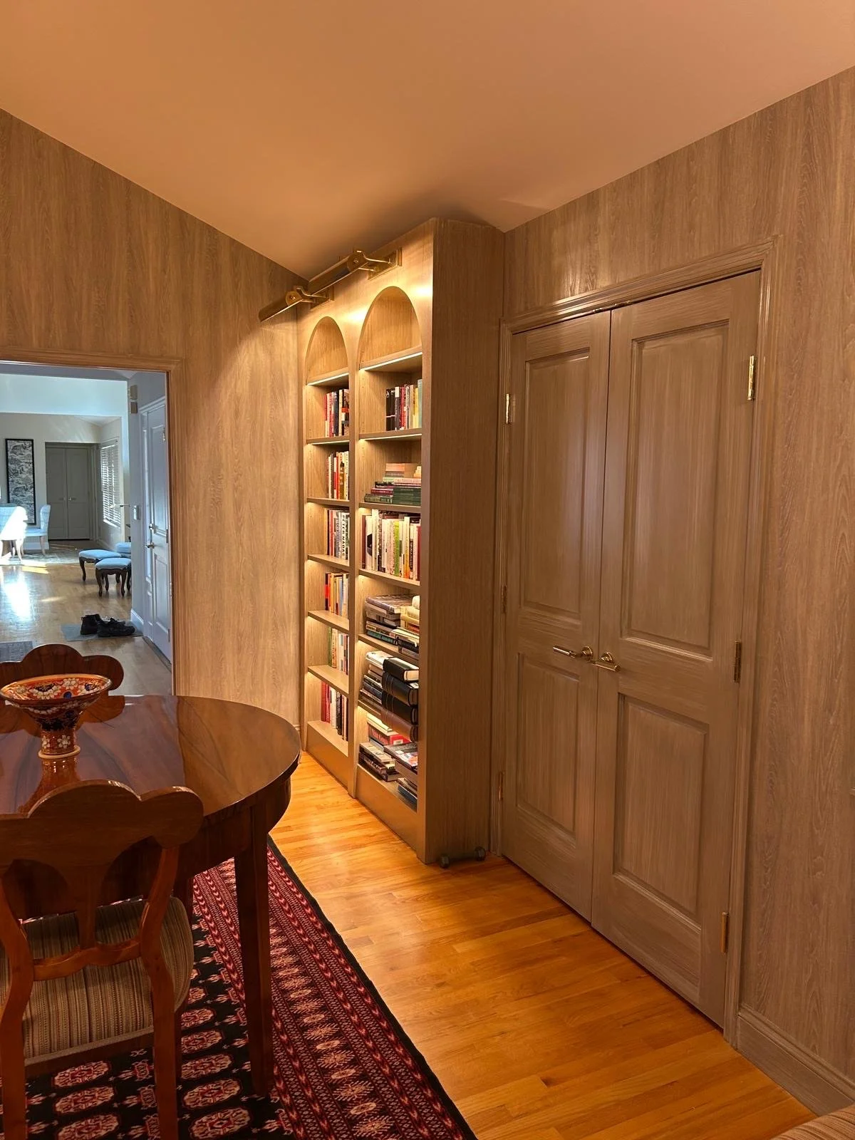 Living room with wood-paneled walls, a bookshelf filled with books, a wooden closet door, a dining table and chairs, and a patterned rug.