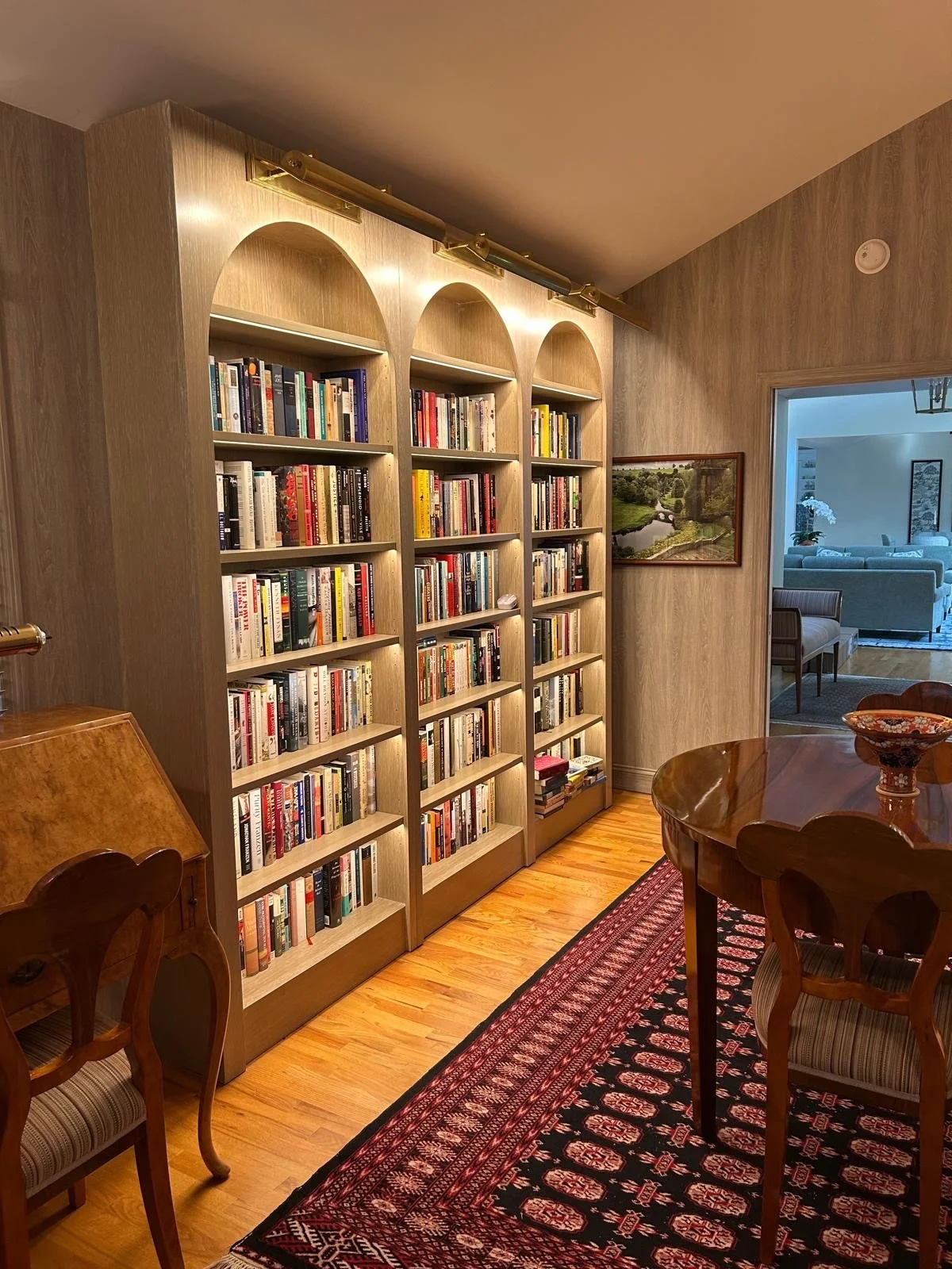 A cozy living room corner with a wooden bookshelf filled with books, a wooden dining table with chairs, and a patterned rug on the hardwood floor. Part of the next room with more furniture is visible through a doorway.