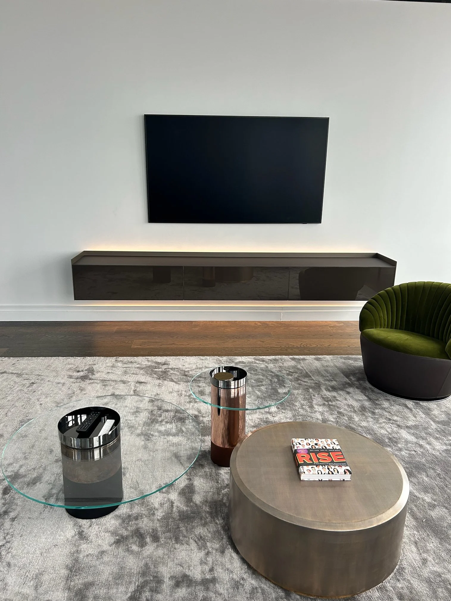 Modern living room with a large flat-screen TV on a white wall, a floating black TV console beneath it, a green velvet swivel chair, two glass coffee tables with wooden bases, a patterned book on one coffee table, and a gray area rug.