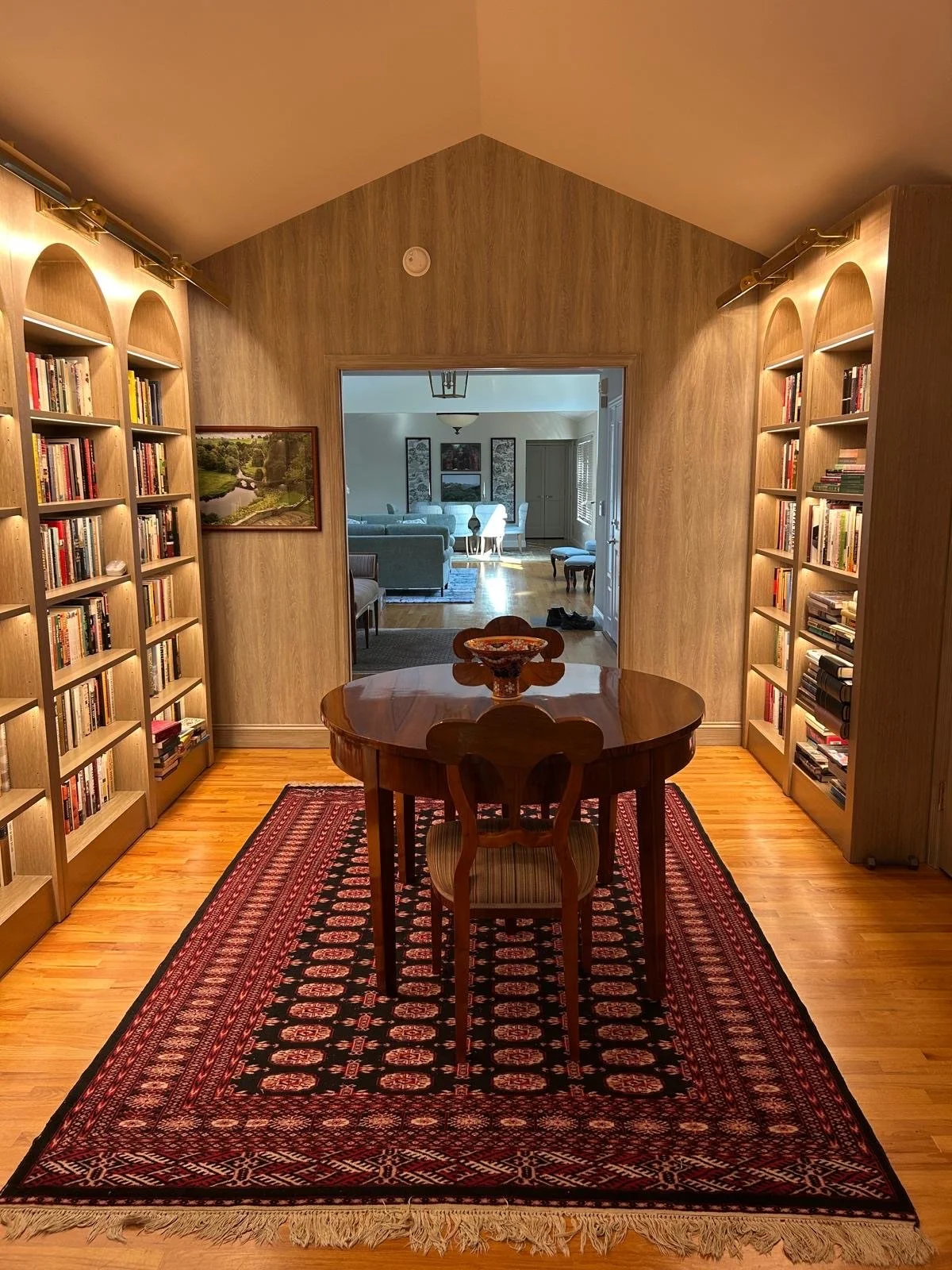 Dining room with a wooden table and a single chair, area rug, bookshelves, and doorway leading to a living room with sofas and artwork.