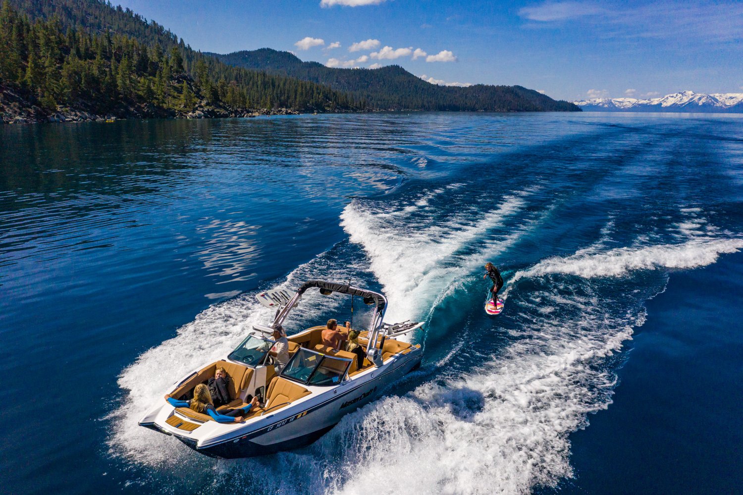 Aerial view of a boat towing a person wakesurfing on a clear blue lake, surrounded by forested mountains.