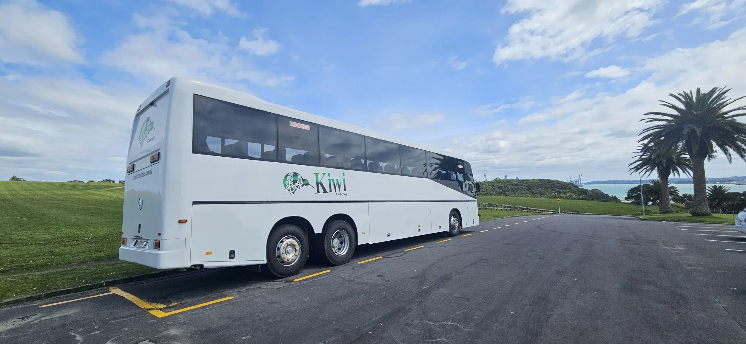 White coach bus with Kiwi Coaches logo parked in a lot near palm trees and green grassy landscape, with a blue sky and water in the background.