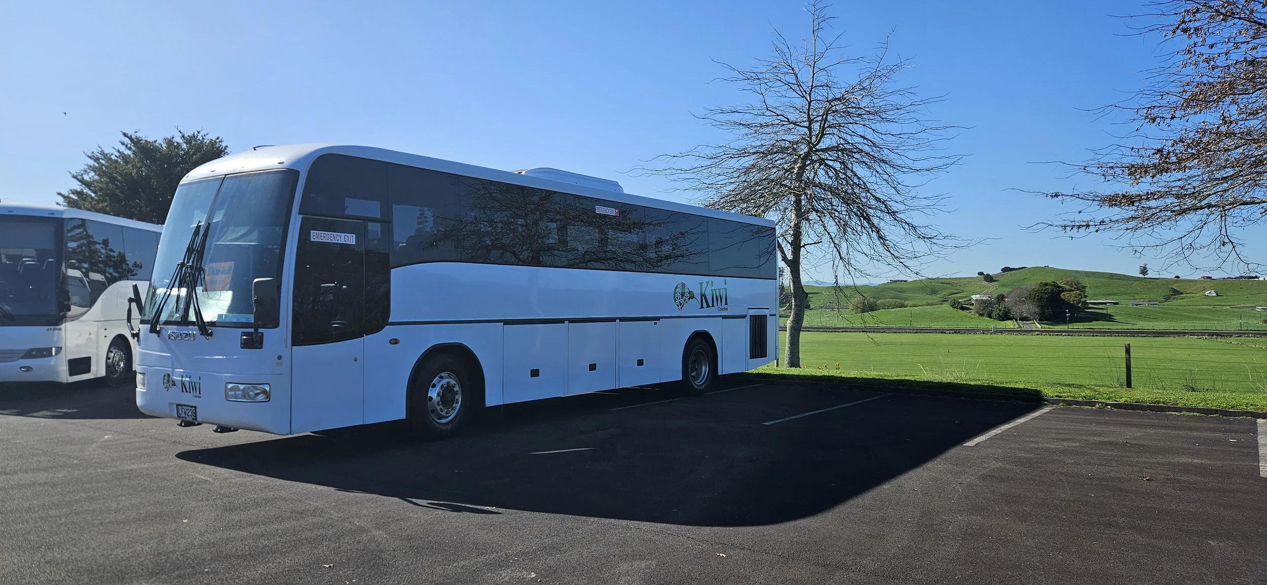 White bus with Kiwi logo parked in a lot with a grassy field and trees in the background on a sunny day.