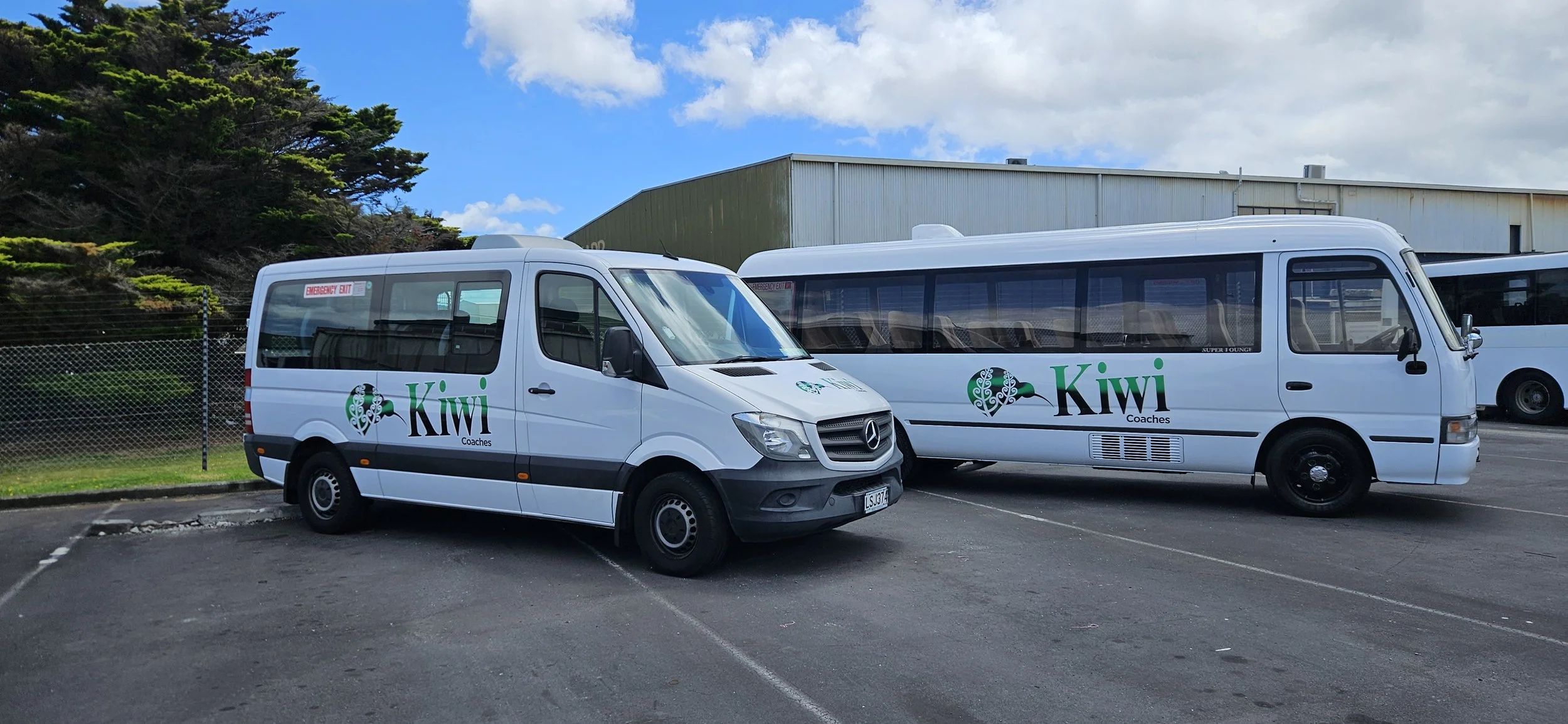 Two white Kia Coaches vans parked on a lot with a chain-link fence and a large industrial building in the background. The vans have the green Kiwi Coaches logo and are under a partly cloudy blue sky.