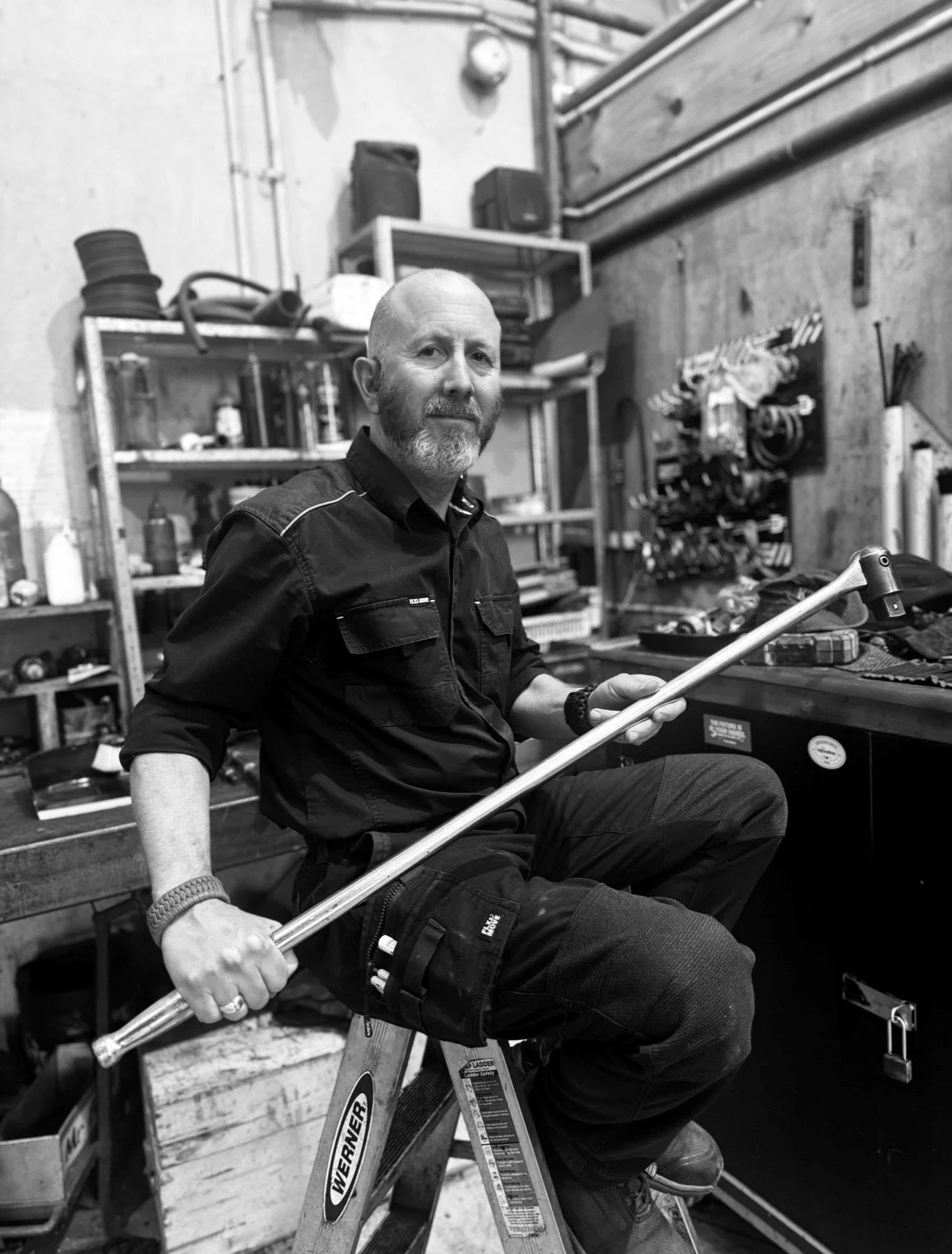 A man with a beard, sitting on a ladder in a workshop, holding a long metal tool, surrounded by various tools and equipment.