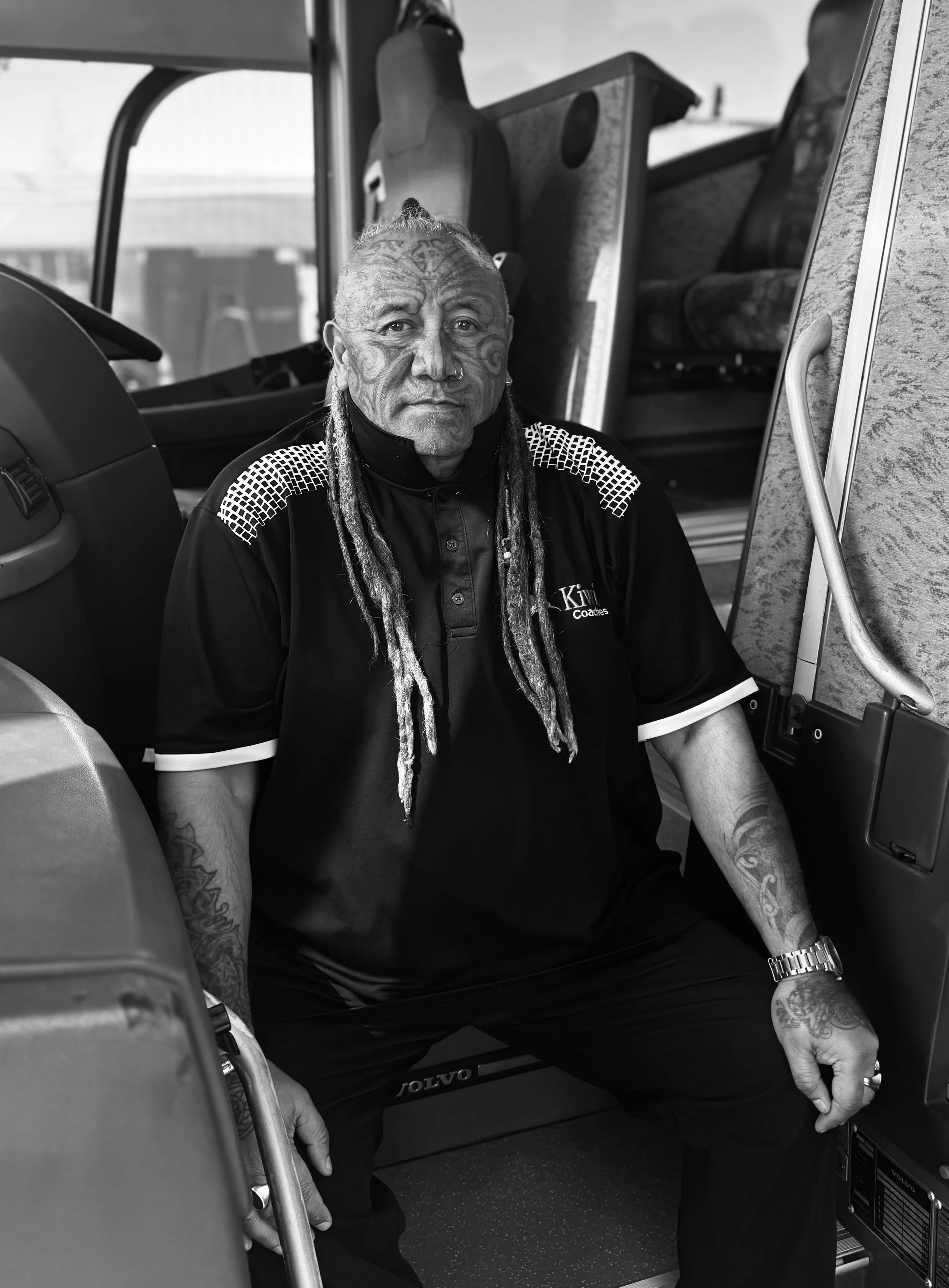 A man with facial tattoos and long dreadlocks sitting inside a bus, wearing a black sports shirt with white accents, with bus seats and a window in the background.