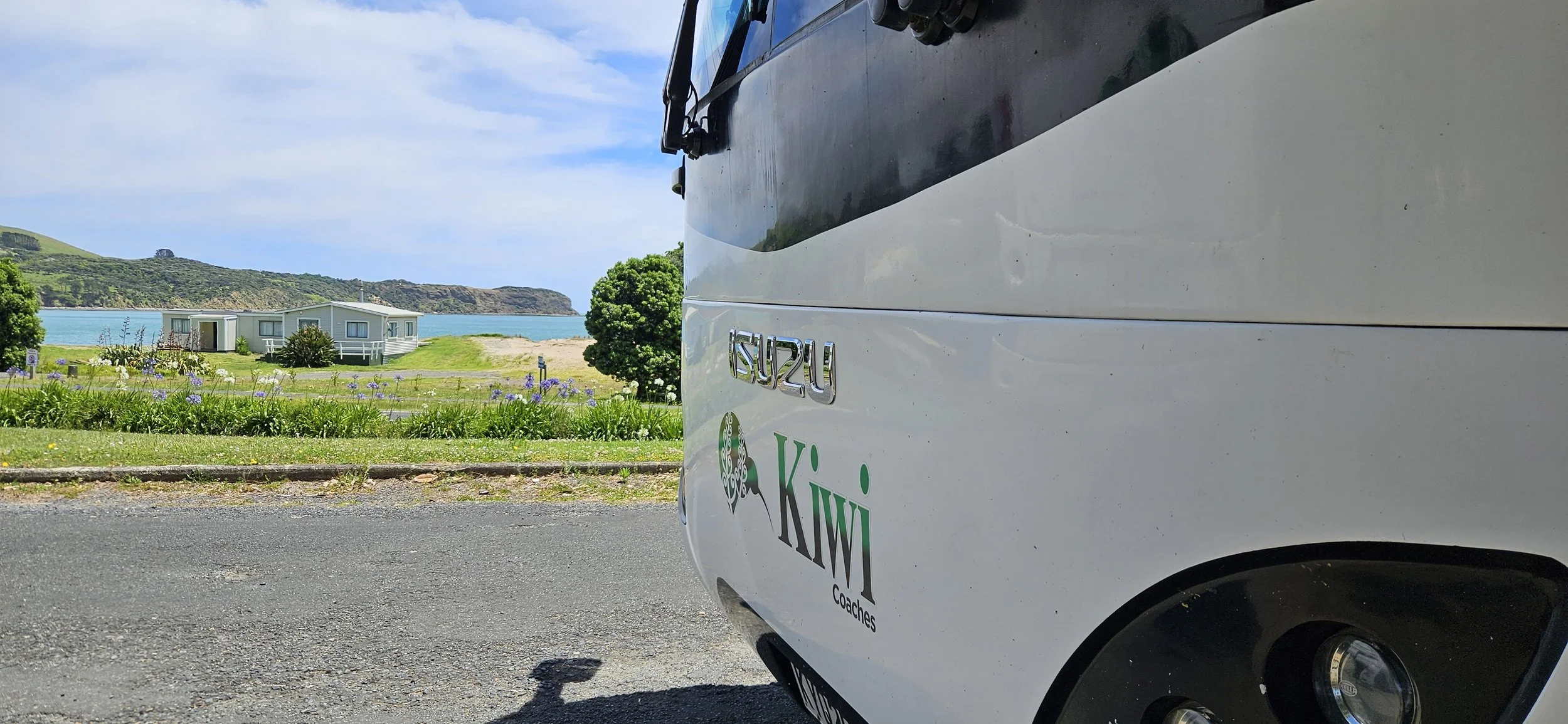 Close-up of a white van with Isuzu logo and Kiwi Coaches branding parked on a street near a coastal area with houses, a grassy field, trees, and hills in the background on a partly cloudy day.