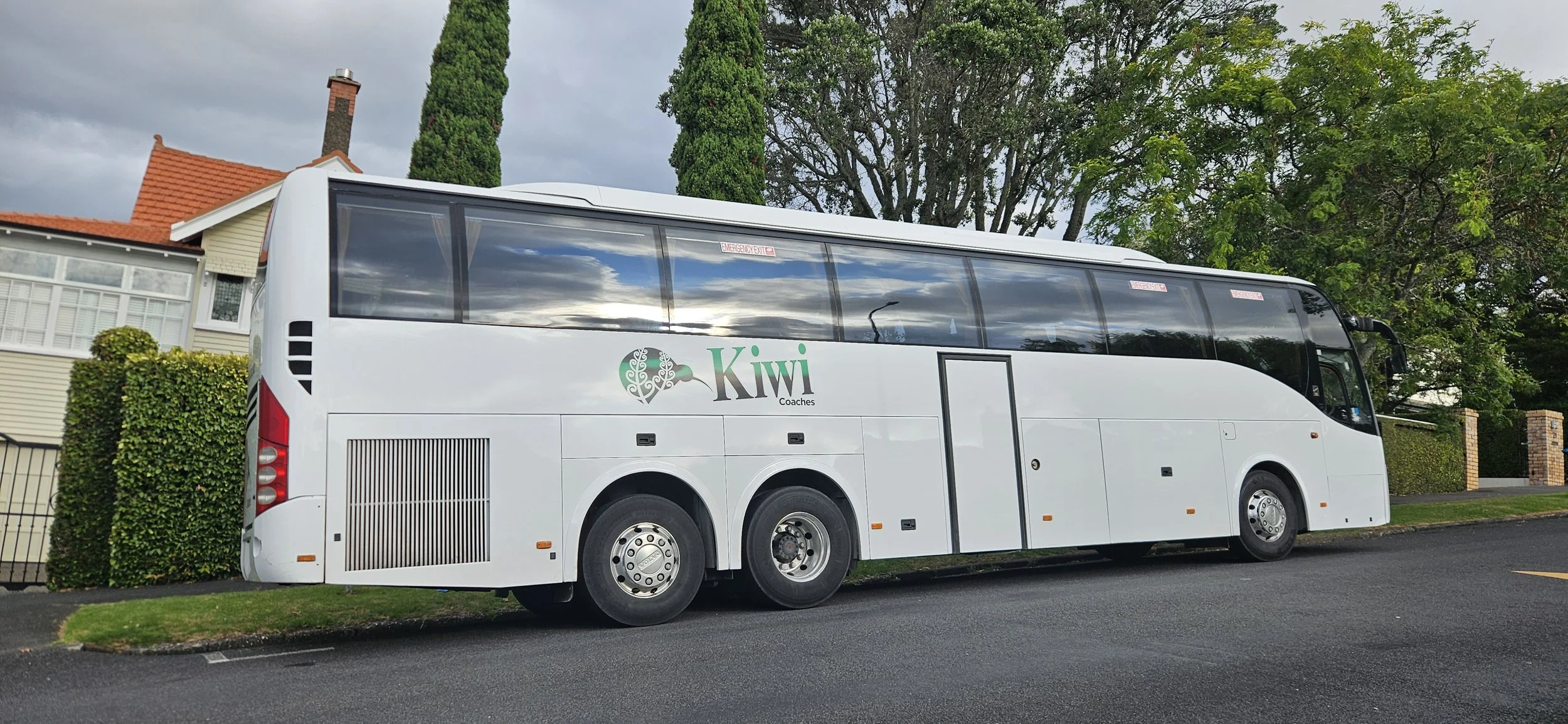 White coach bus labeled 'Kiwi Coaches' parked on a street in front of houses and trees.