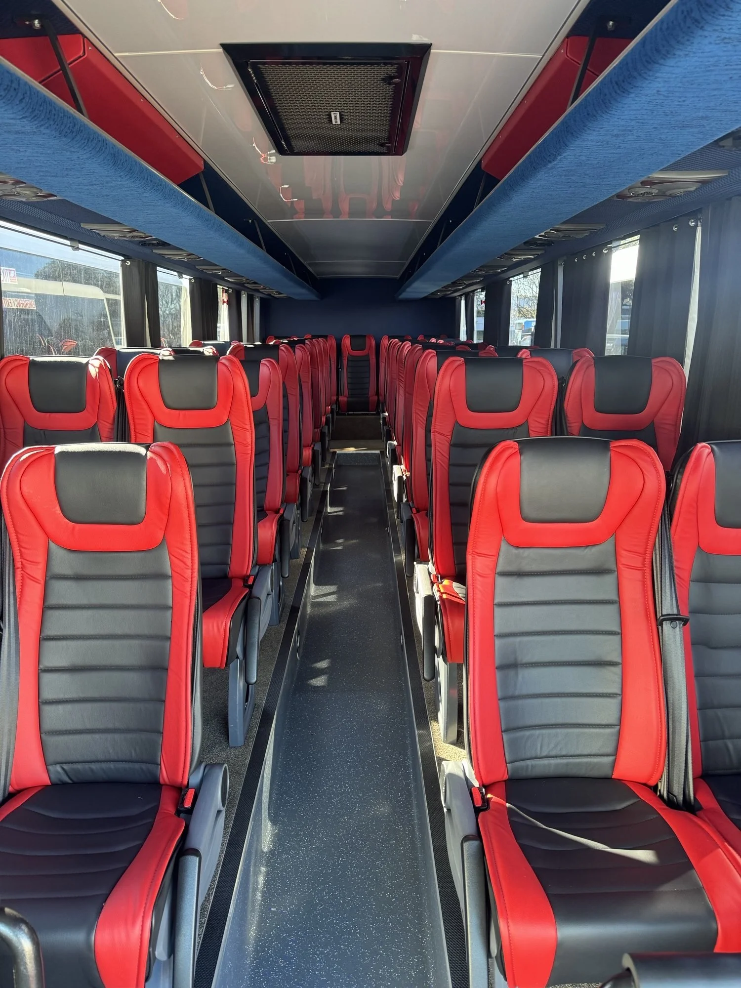 Interior of a modern bus with red and black seats, aisle in the middle, windows on both sides, and overhead storage compartments.