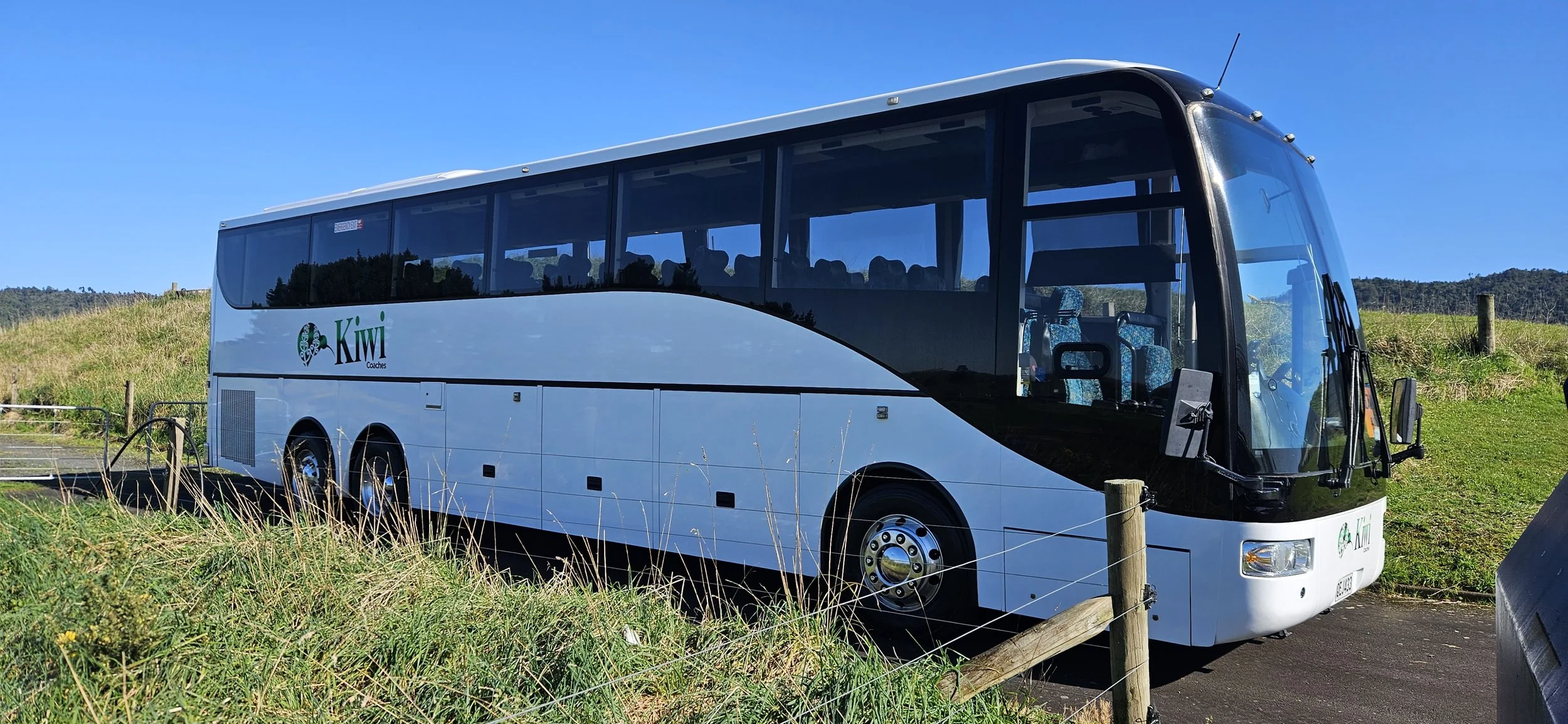 A white bus with black accents, labeled 'Kiwi Coaches,' parked on the side of a rural road with grassy fields and hills in the background under a clear blue sky.