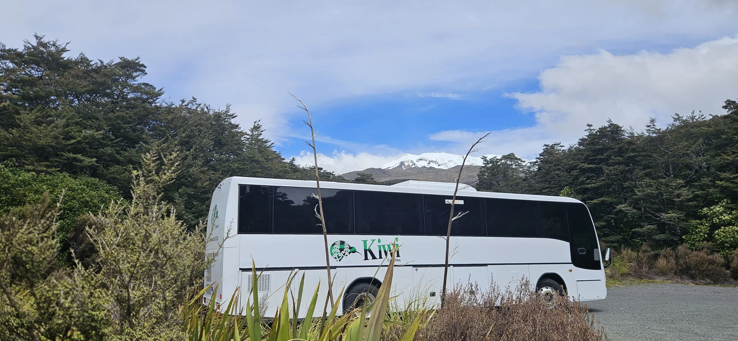 White tour bus with 'Kiwi' logo parked on a gravel area surrounded by green shrubs and trees, with snow-capped mountains and partly cloudy sky in the background.
