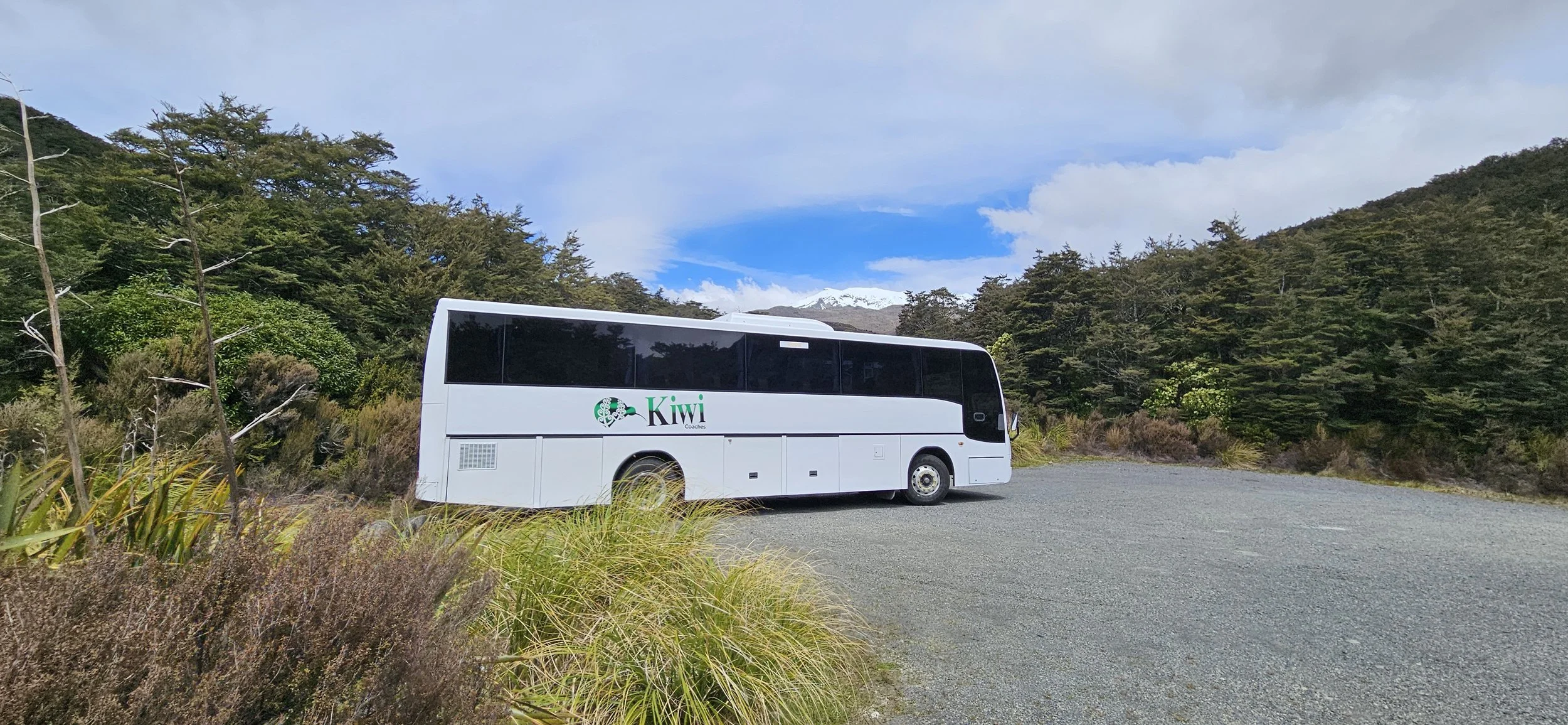 A white bus with 'Kiwi Coaches' logo parked on a gravel area surrounded by greenery and trees, with mountains and partly cloudy sky in the background.