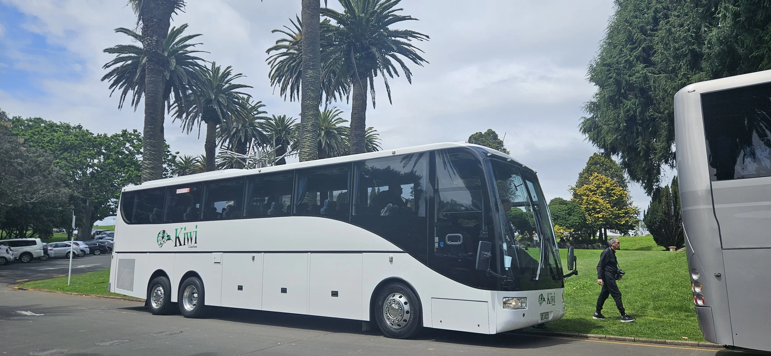 A white tour bus parked on a street with palm trees and green grass in the background. A person is walking on the grass near the bus, holding a camera.