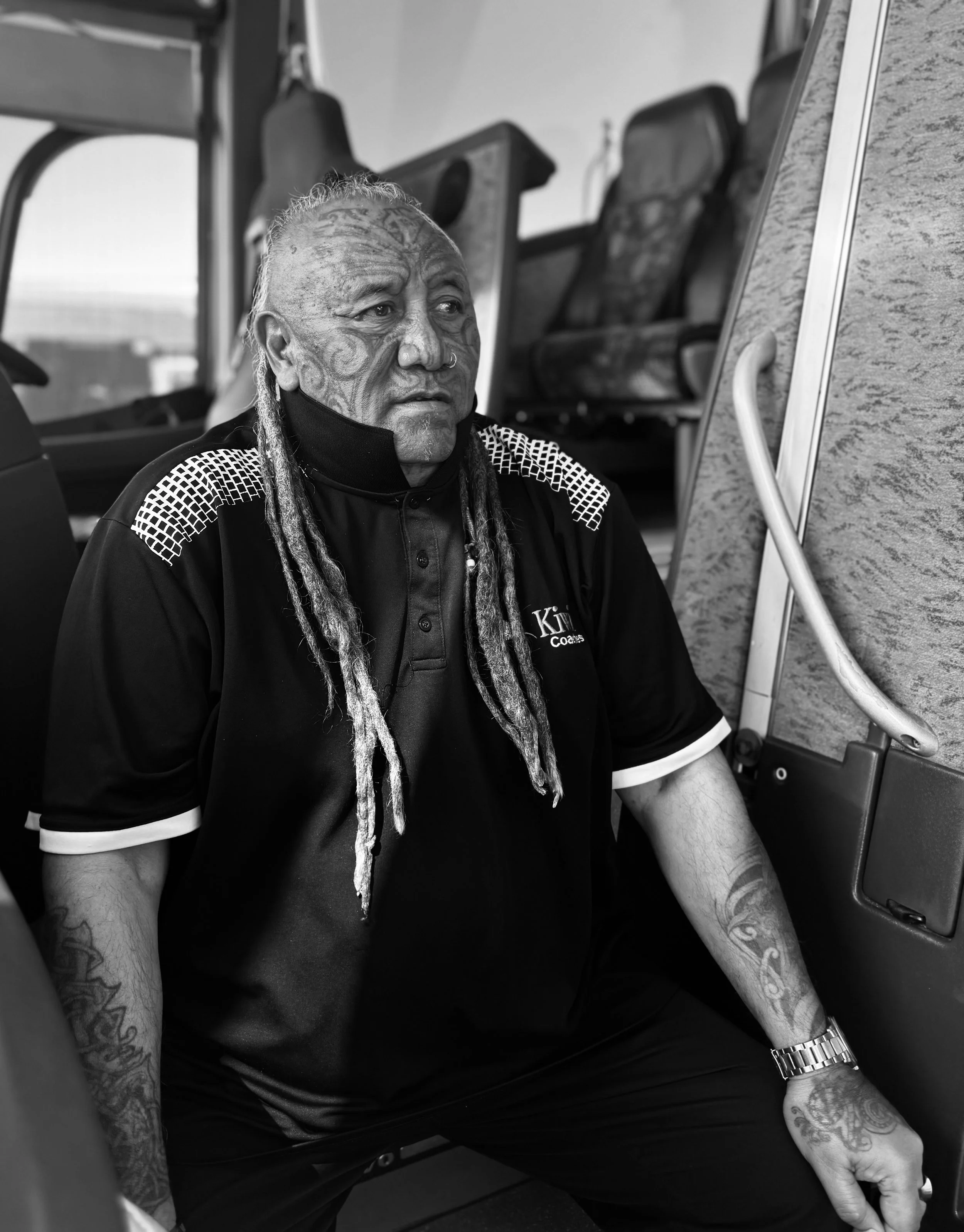 A tattooed man with long dreadlocks sitting on a bus, looking serious, wearing a black collared shirt with white accents, and a wristwatch, with bus seats and window in the background.