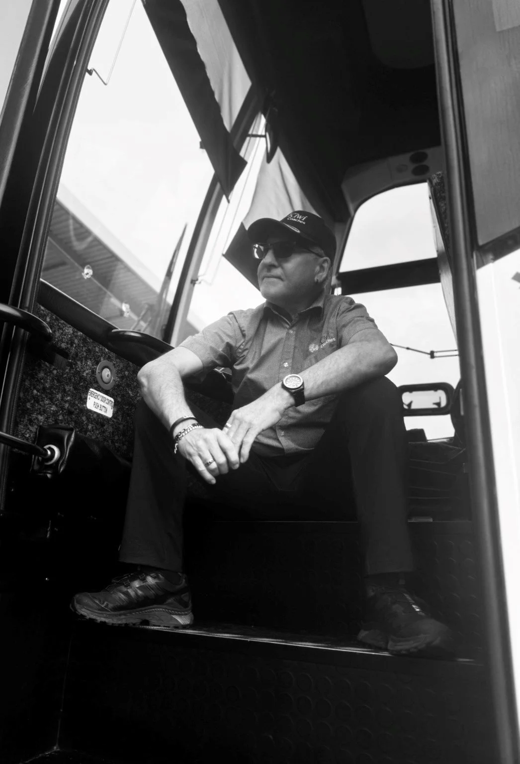 A man sitting inside a forklift, wearing sunglasses, a cap, a watch, and a bracelet, with his hands resting on his knee.