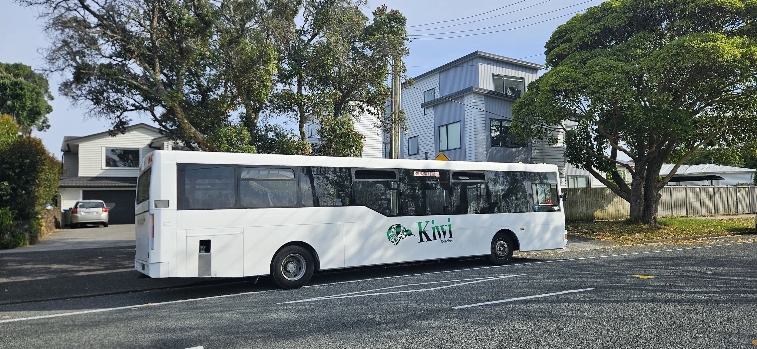 A white bus with 'Kiwi Coaches' logo parked on a city street near residential houses and trees.